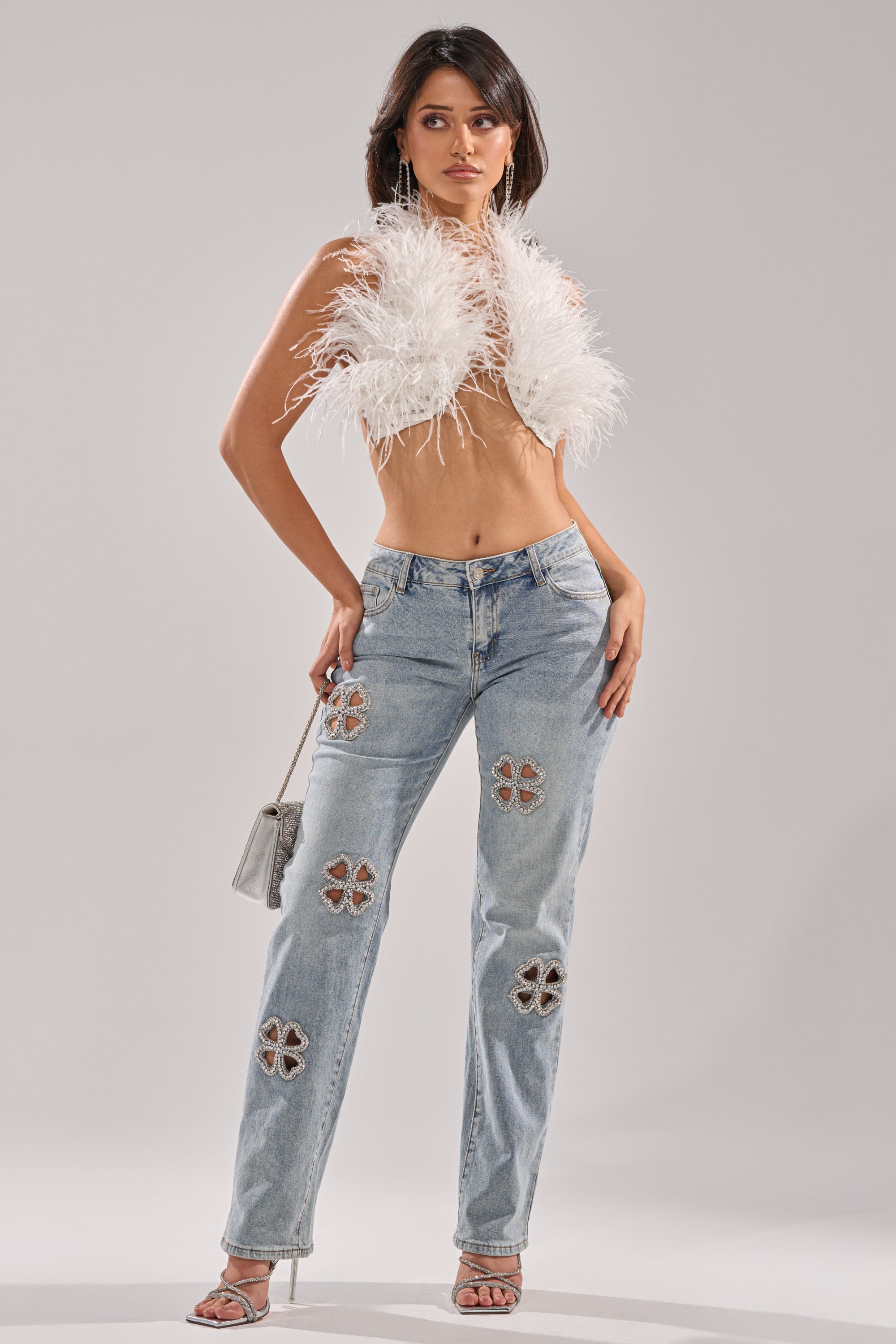A woman stands confidently in DIVINE FEMININE ENERGY RHINESTONE DETAIL JEANS, a white feathered crop top, high heels, and a small silver handbag against a plain gray background.