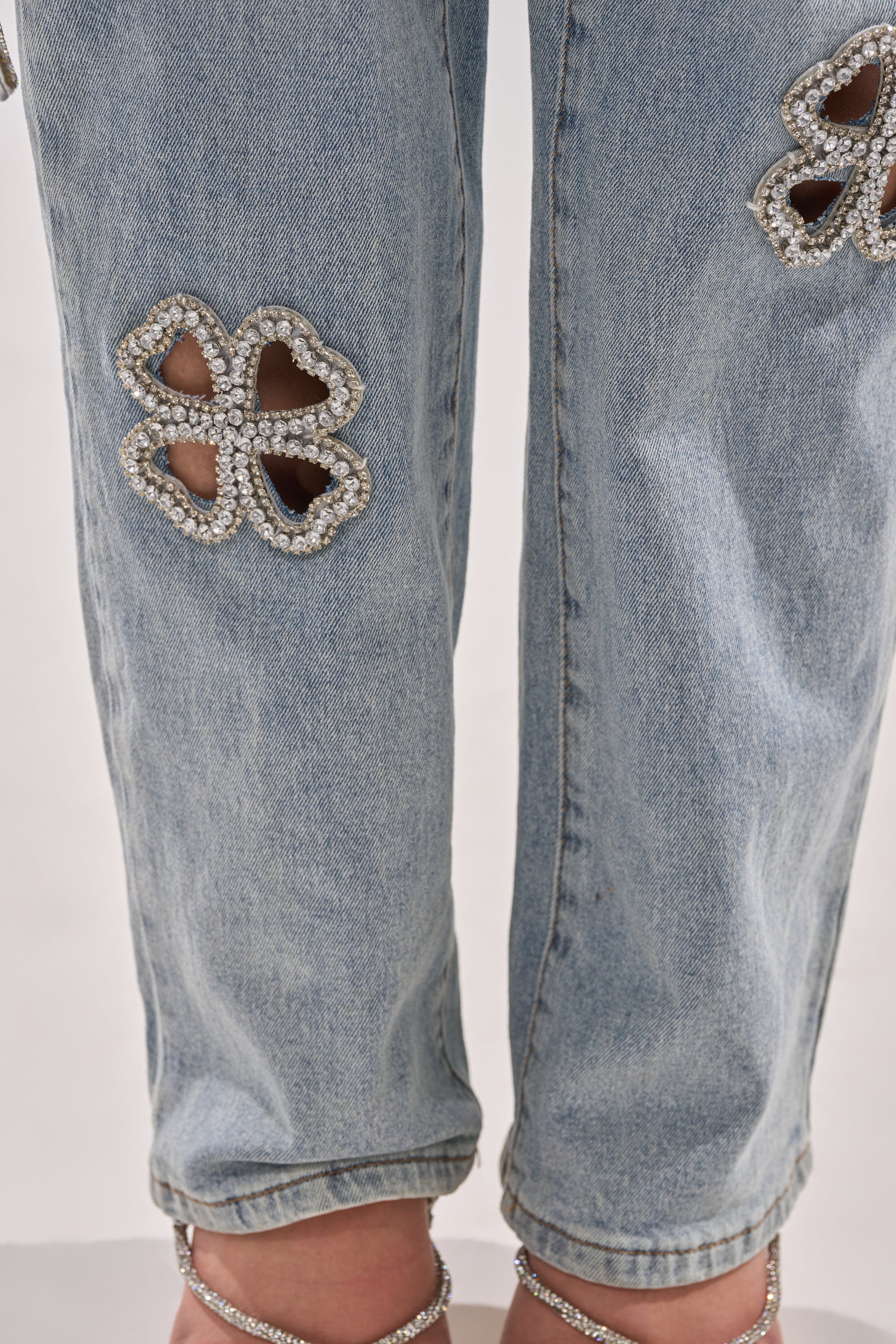 Close-up of DIVINE FEMININE ENERGY RHINESTONE DETAIL JEANS in light wash denim with flower-shaped rhinestone-outlined cutouts at the knees. The hems are slightly rolled, and open-toe heels peek out at the bottom.
