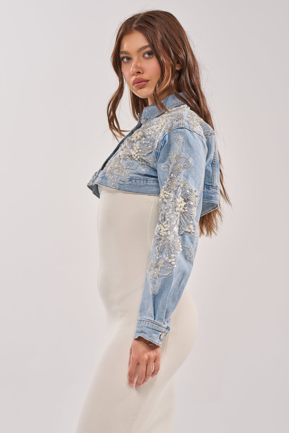 A woman with long brown hair stands sideways against a plain background, wearing the BILLIONAIRE BELLE EMBELLISHED DENIM JACKET with intricate beadwork over a fitted, cream-colored dress. 3