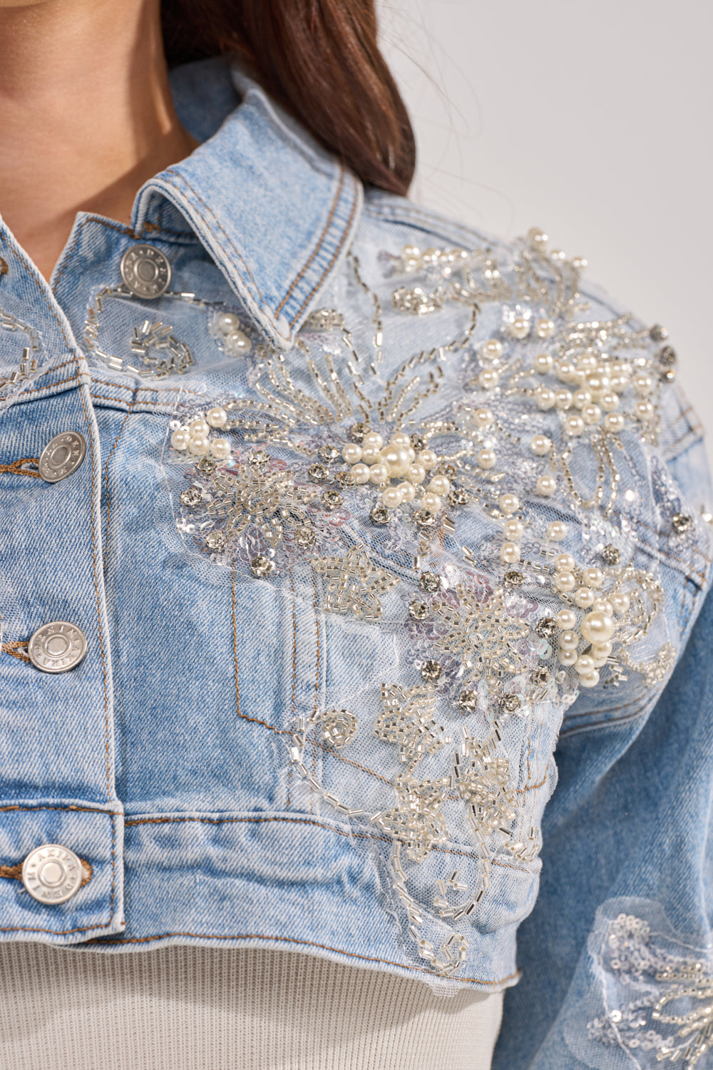 Close-up of the BILLIONAIRE BELLE EMBELLISHED DENIM JACKET, featuring intricate beadwork, pearls, and silver details on the shoulder and chest. The cropped denim jacket is styled with a white top, partially revealing the wearer’s neck and hair. 7