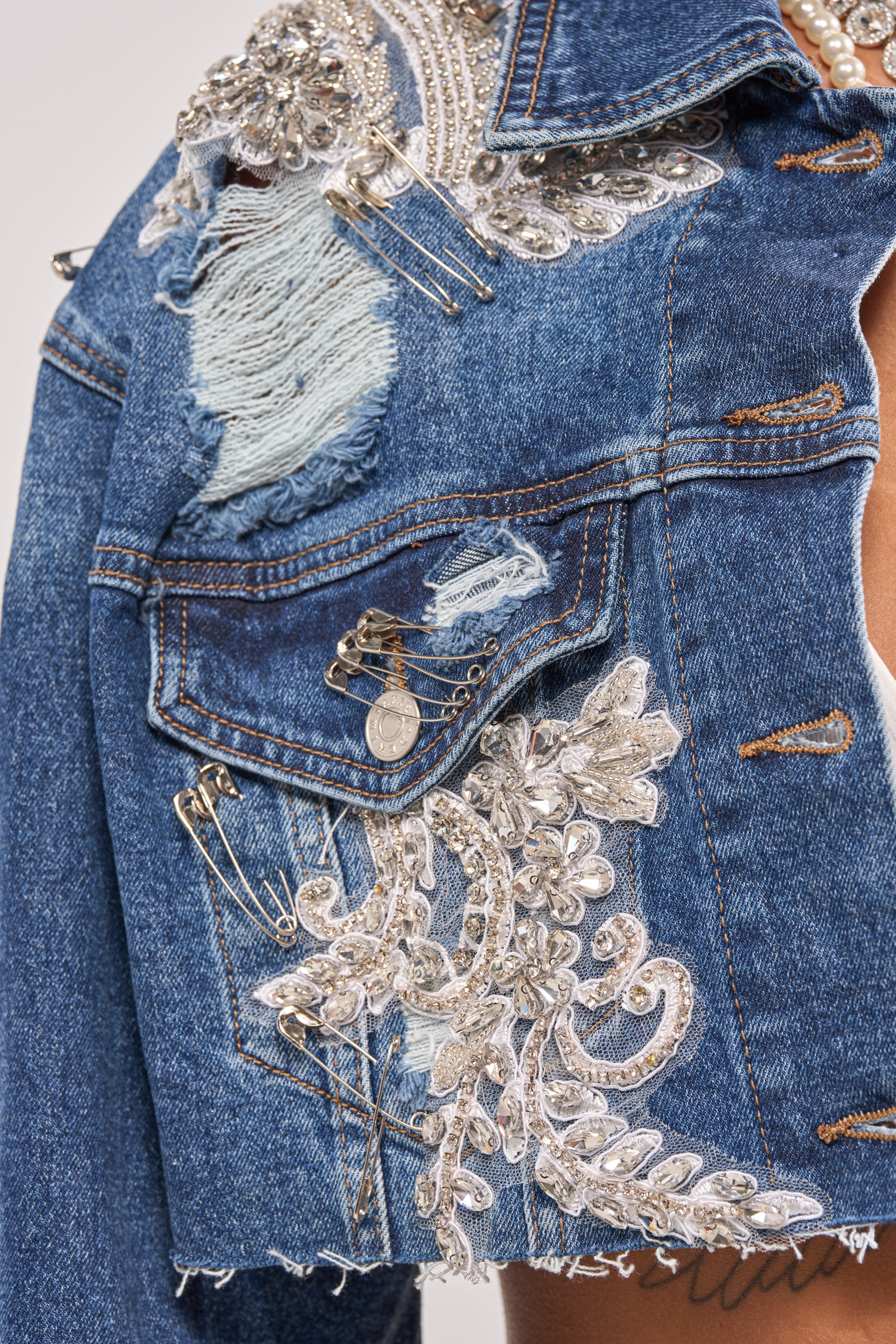 Close-up of the LET'S RIDE EMBELLISHED DENIM JACKET: a cropped blue denim jacket featuring silver sequins, beads, embroidered florals, safety pins, a distressed patch above the pocket, visible stitches, and frayed edges for standout style. 10