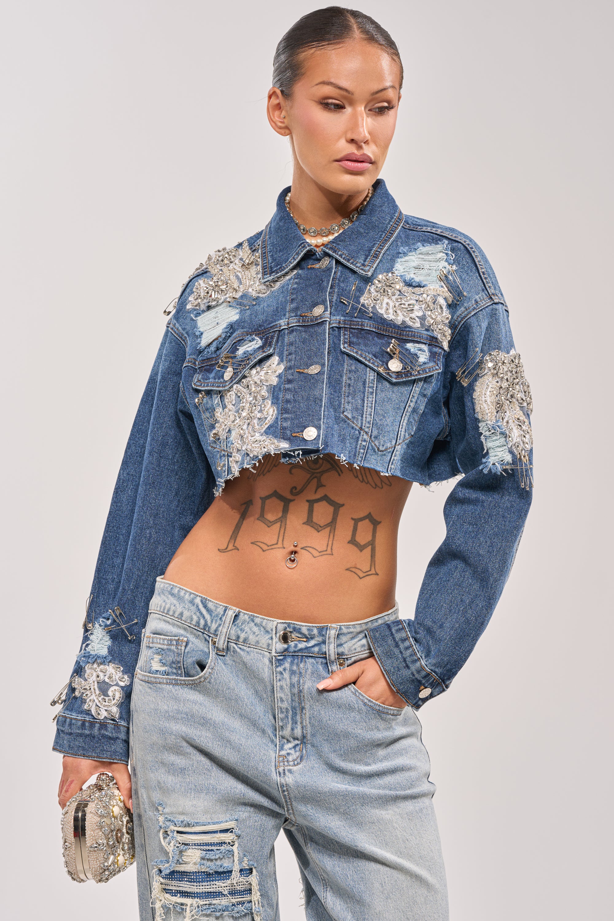 A woman stands with one hand in her pocket, showing off her "1999" tattoo while wearing the LET'S RIDE EMBELLISHED DENIM JACKET and distressed jeans, holding a sparkly purse against a plain background. 1