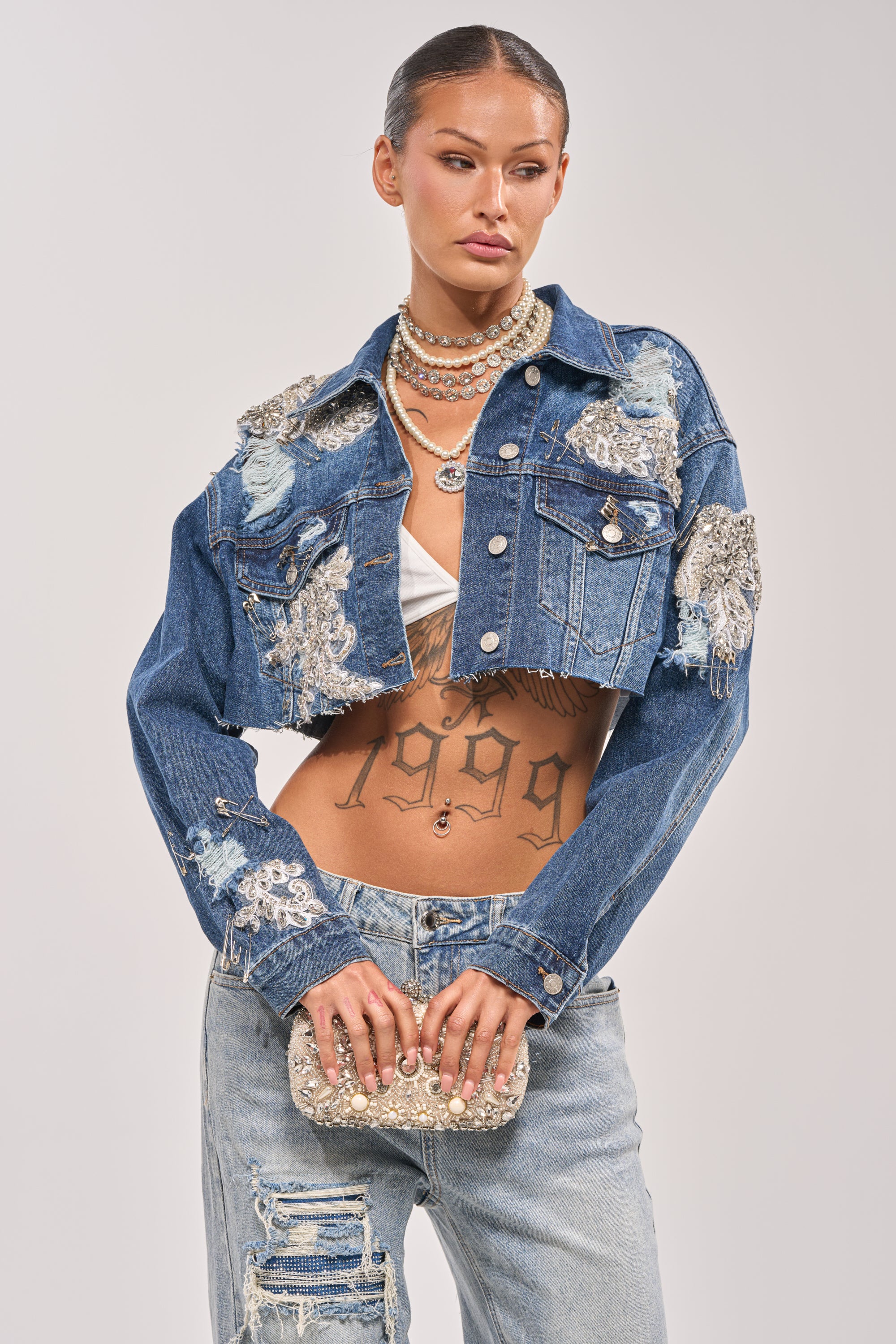 A woman styles the LET'S RIDE EMBELLISHED DENIM JACKET—rhinestone-adorned and cropped—over a white bra top with light blue ripped jeans, layered necklaces, a sparkling clutch, visible torso tattoos, and slicked-back hair. 2