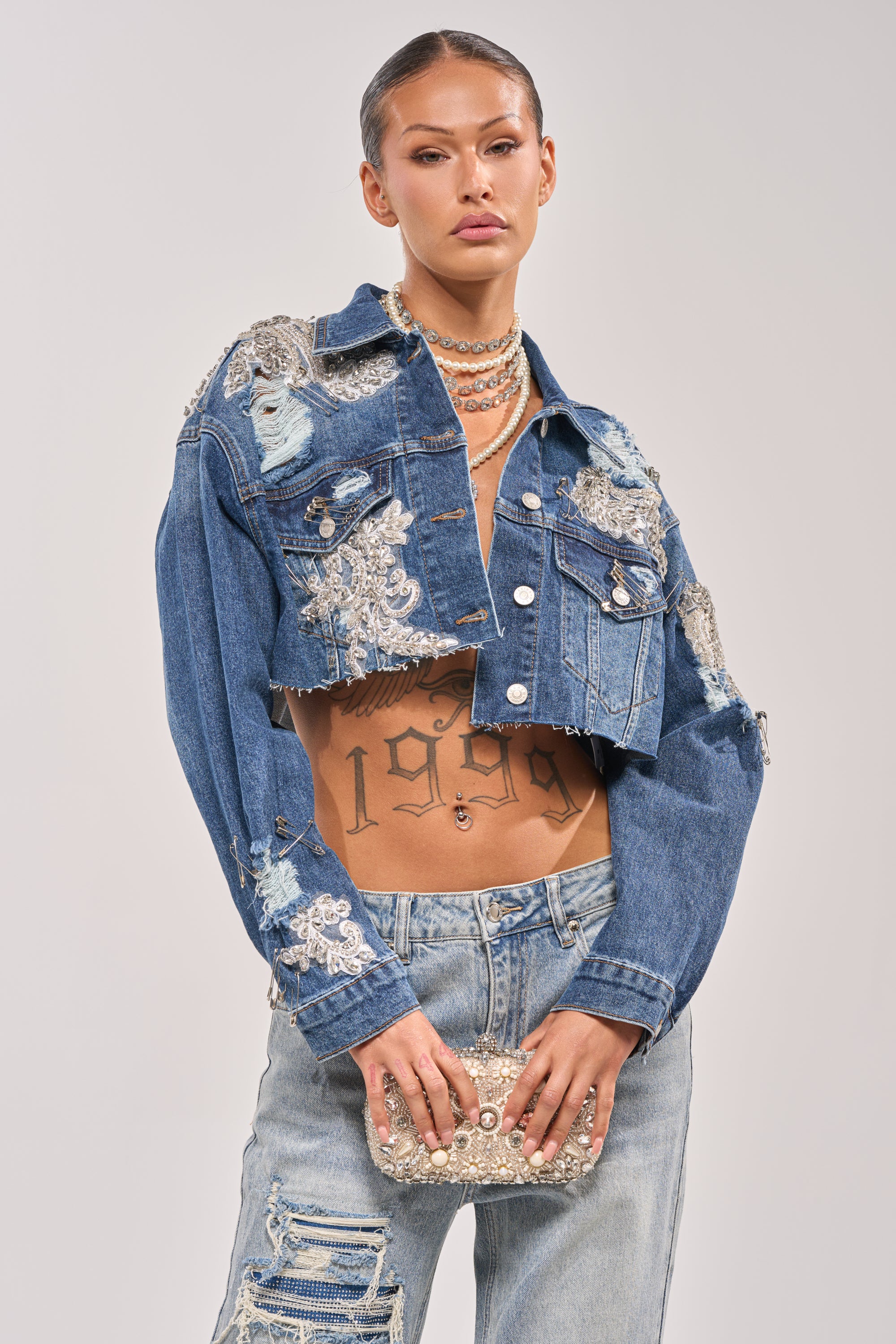 Wearing the LET'S RIDE EMBELLISHED DENIM JACKET, distressed jeans, layered necklaces, and holding a beaded clutch, the person shows off a midriff tattoo reading "1996" beneath clusters of rhinestones. 3