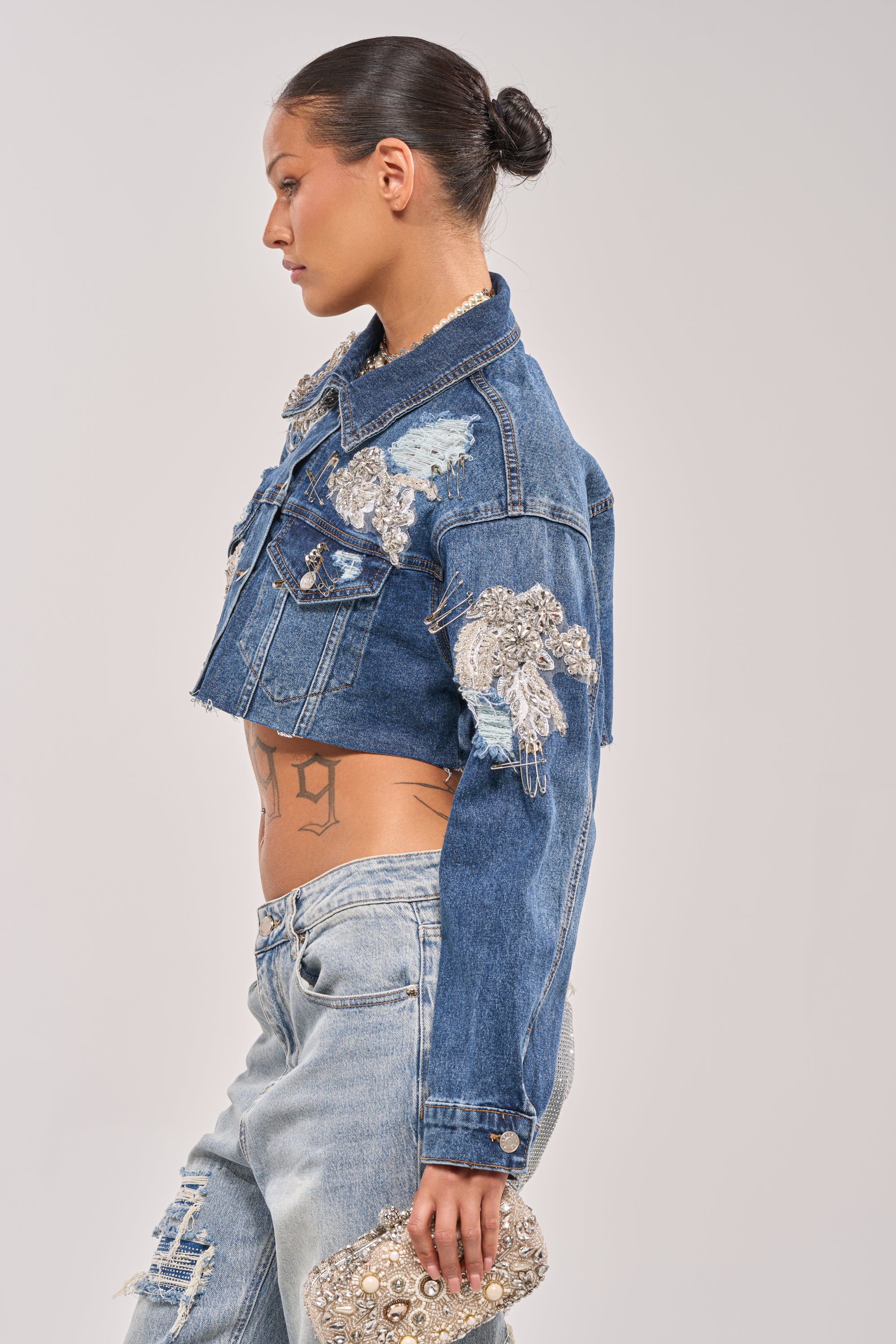 Woman in profile wears the LET'S RIDE EMBELLISHED DENIM JACKET with sequins and patches, paired with light ripped jeans and a beaded clutch, her hair in a neat bun showing a geometric side tattoo. 5