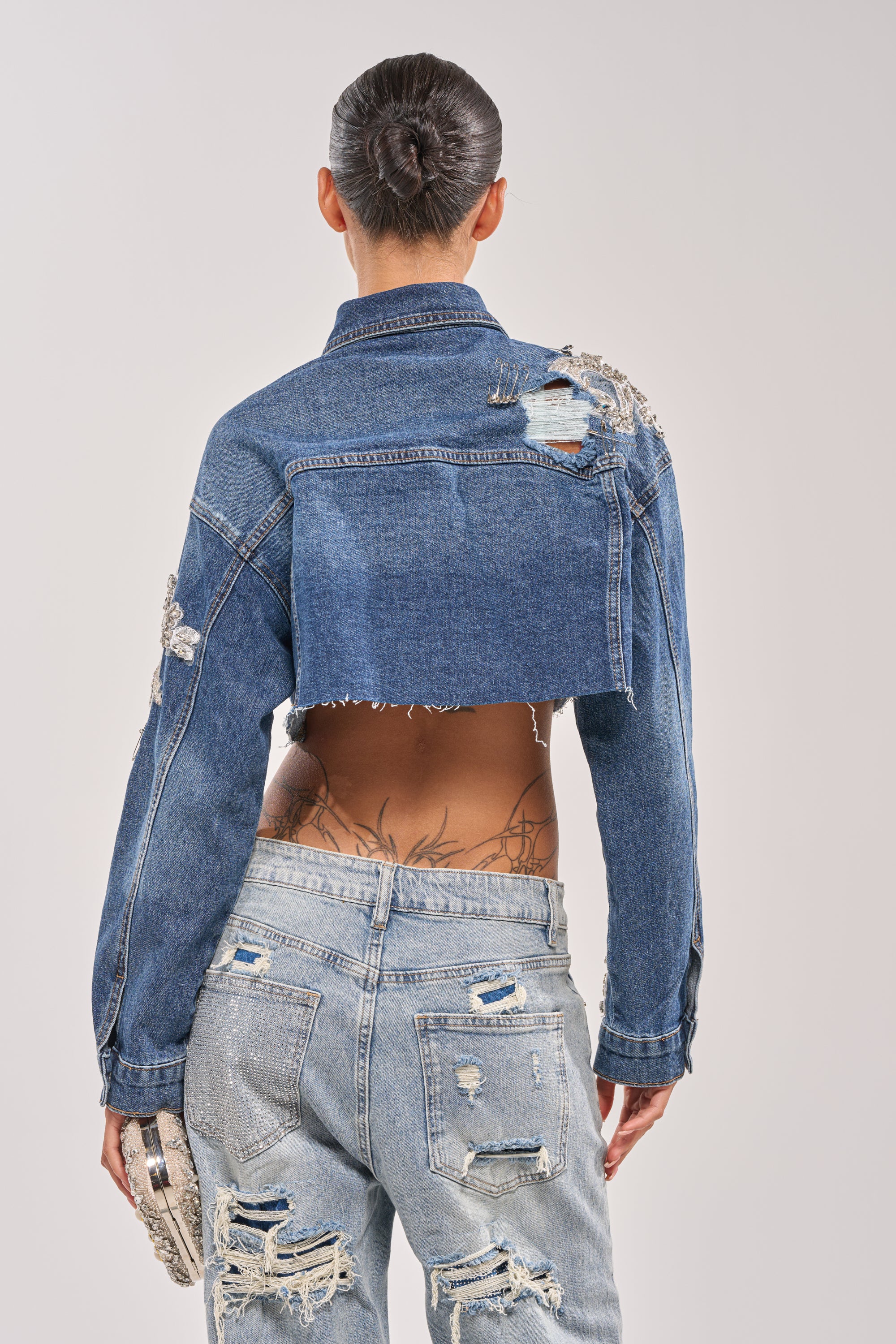 Seen from behind, a person with hair in a sleek bun wears the LET'S RIDE EMBELLISHED DENIM JACKET styled cropped, with ripped light-wash jeans. A lower back tattoo and small textured clutch complete their trendy look. 6