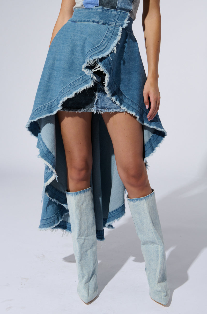 A person is wearing a blue denim high-low skirt with a raw, frayed hem design. The skirt, similar to the Kelsey Overlay Wrap Skirt Belt, pairs perfectly with tall, light-wash denim boots. The background is plain and light-colored. The person's upper body is not fully visible in the image.