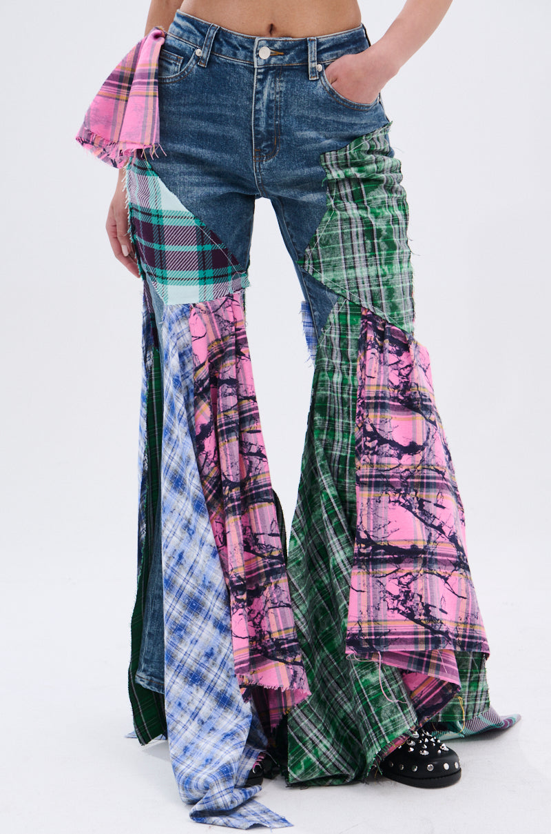 A person models the PLAID ON PLAID DENIM JEANS—wide-leg denim with dramatic flares, frayed edges, and mismatched patchwork in pink, green, blue, and purple plaid patterns.