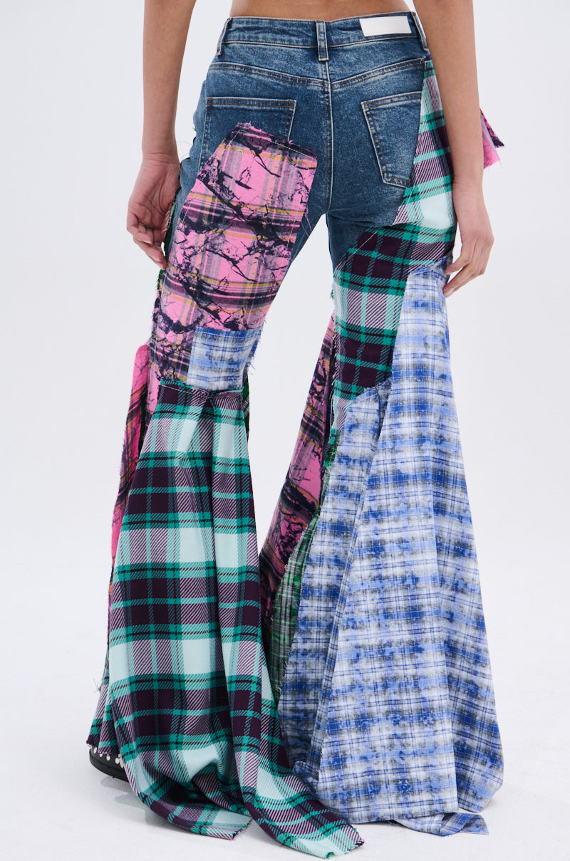 A person wears PLAID ON PLAID DENIM JEANS with wide, flared bell-bottoms made of colorful patchwork plaid and tie-dye fabrics, shown from the back against a plain background.