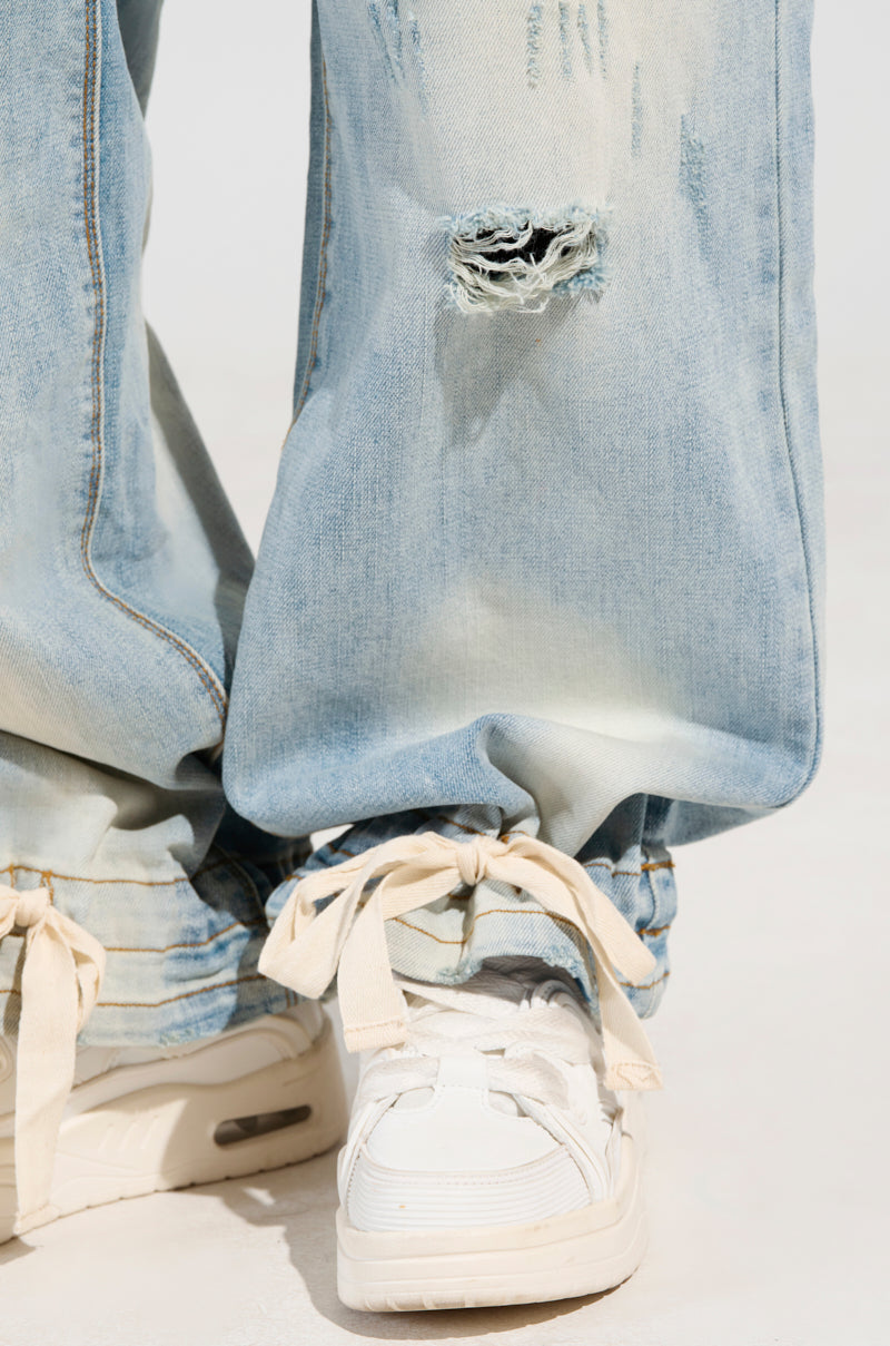 A close-up of a person wearing FOREVER AND A YEAR DENIM JEANS, featuring a light blue hue with a high-rise fit and wide-leg silhouette, paired with white sneakers. The hem of the jeans is slightly frayed and rests on the shoes with loosely tied bows against a plain white background.