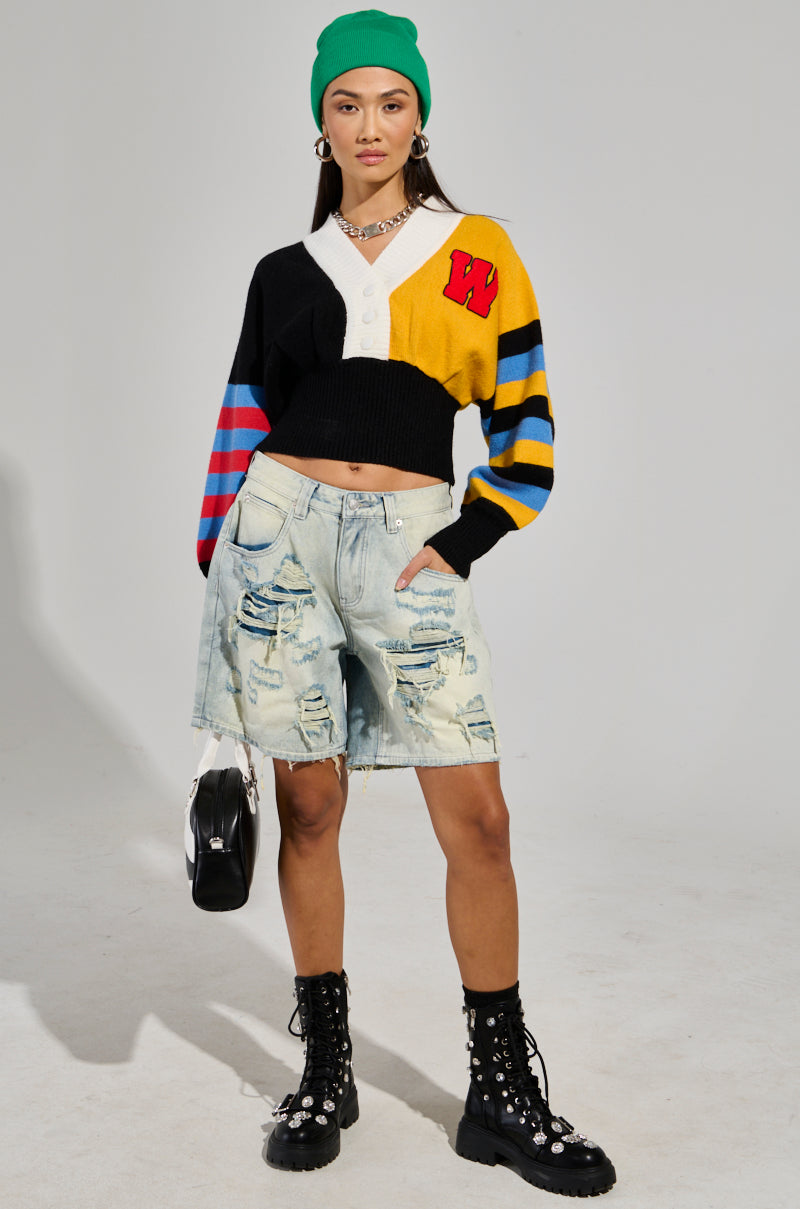 A person wearing a vibrant, striped cardigan featuring the letter "W," paired with LITE UP DENIM SHORTs, black studded boots, a green beanie, and a necklace poses confidently. They are holding a small black bag and stand against a plain background.