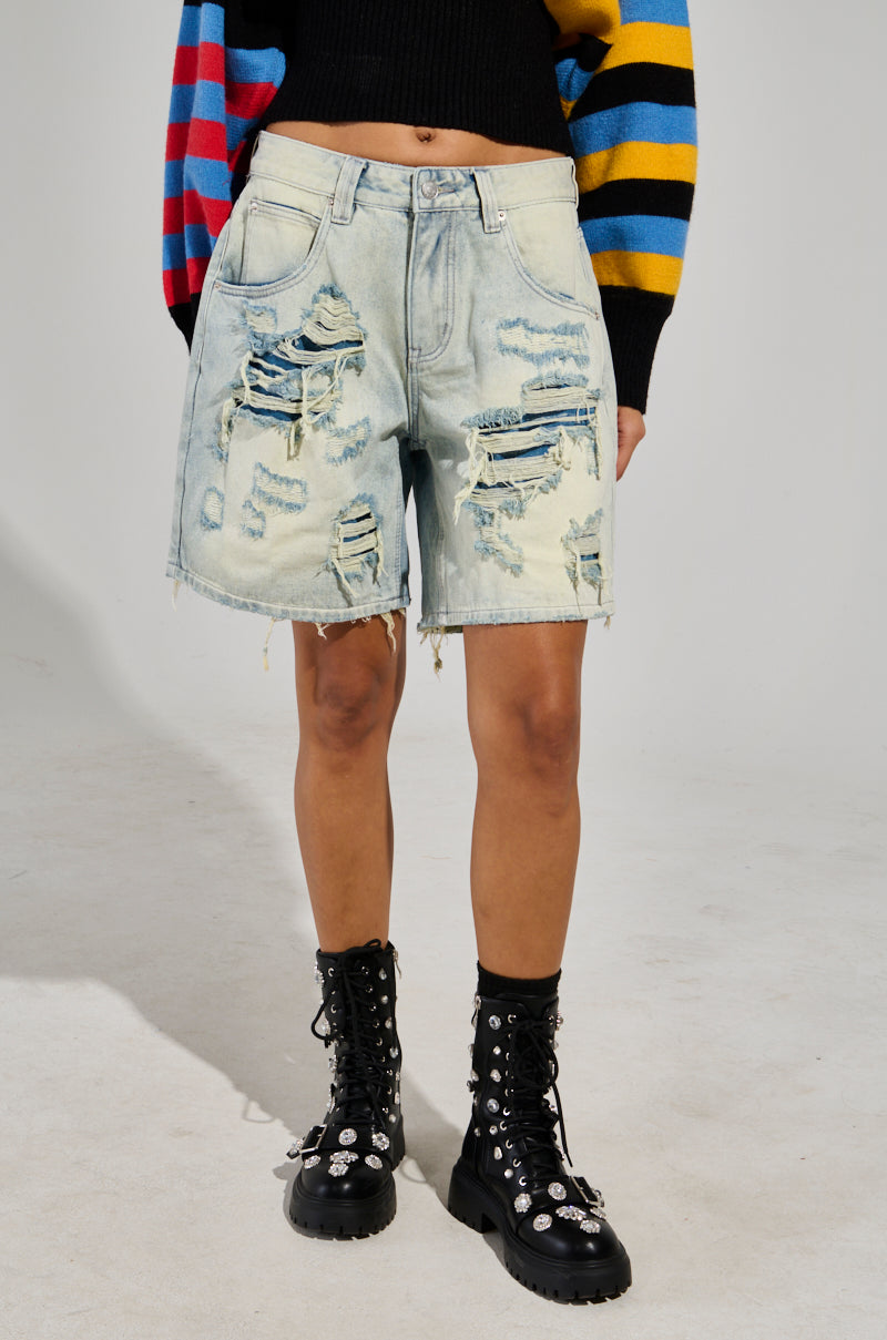 A person wearing LITE UP Denim Shorts, black lace-up boots with decorative elements, and a colorful striped sweater. The background is plain and light-colored.