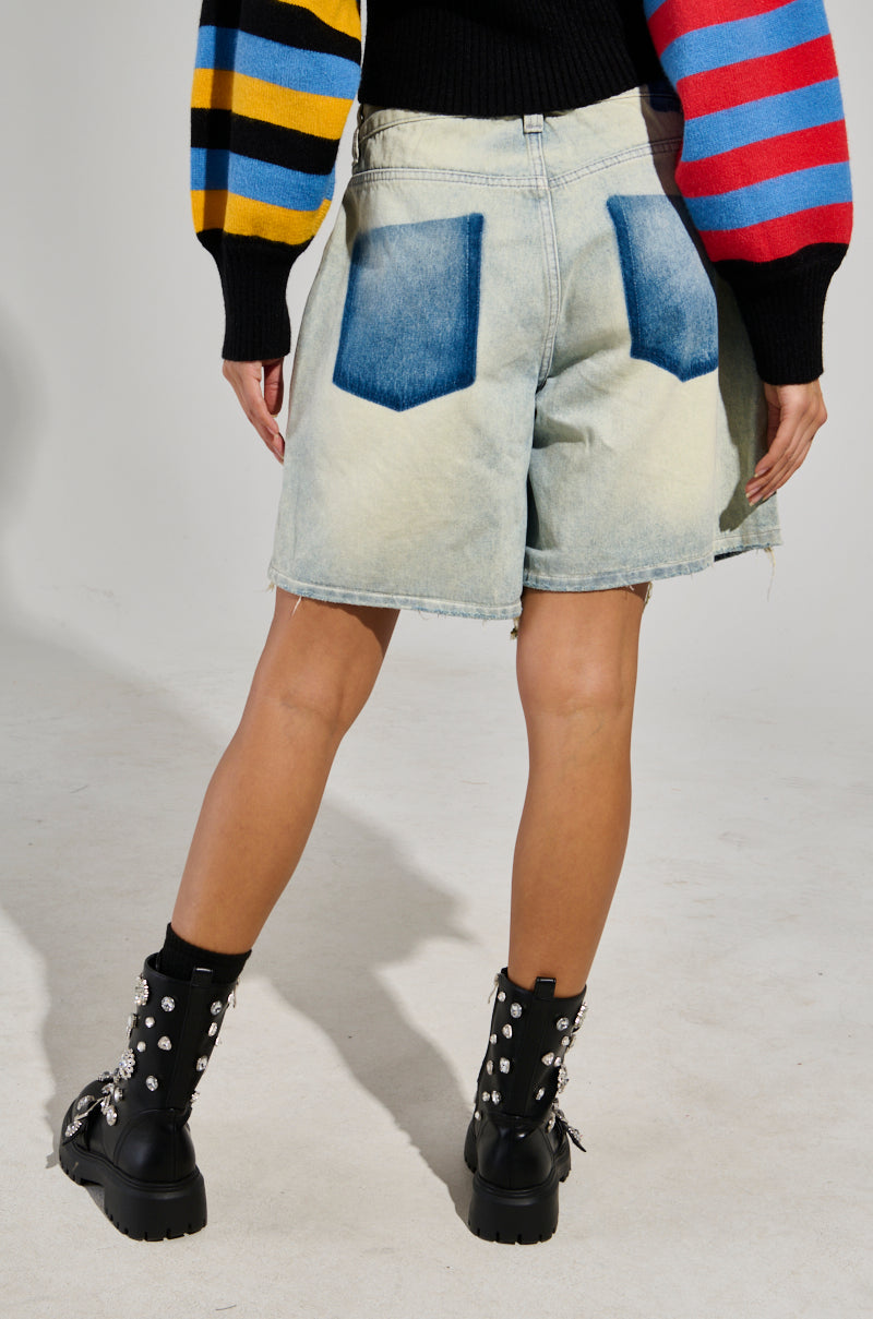 A person stands with their back turned, showcasing the LITE UP DENIM SHORT in light blue. The high-rise shorts are accented by distinct dark blue back pockets and distressed detailing, paired with a vibrant striped sweater. They complete the look with black boots embellished with silver studs against a simple light gray backdrop.