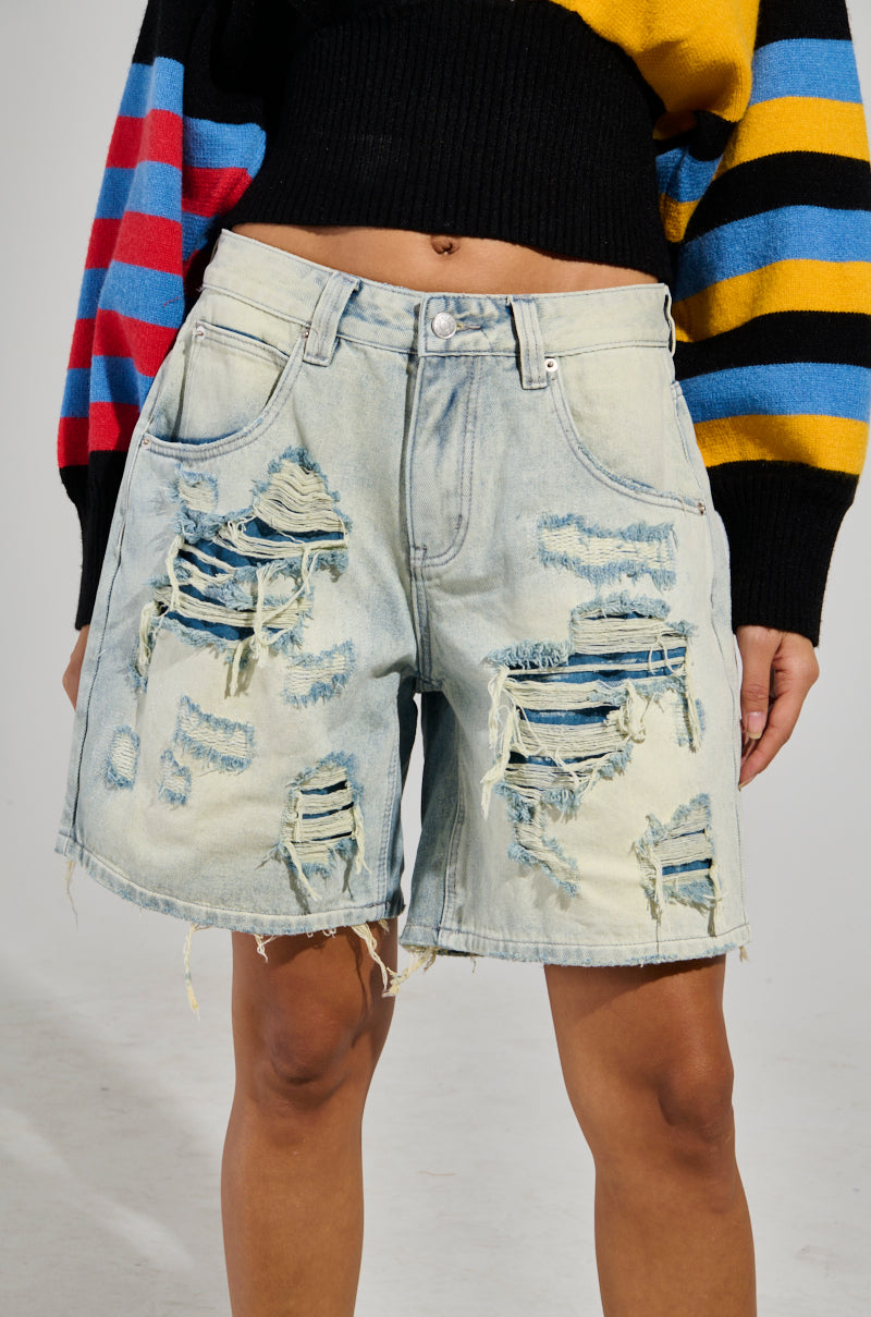 A person is sporting the LITE UP DENIM SHORT, characterized by a high-rise fit and light wash with distressed detailing and multiple rips that reveal a layer of dark blue stripes beneath. Additionally, the individual is wearing a multicolored, striped sweater.