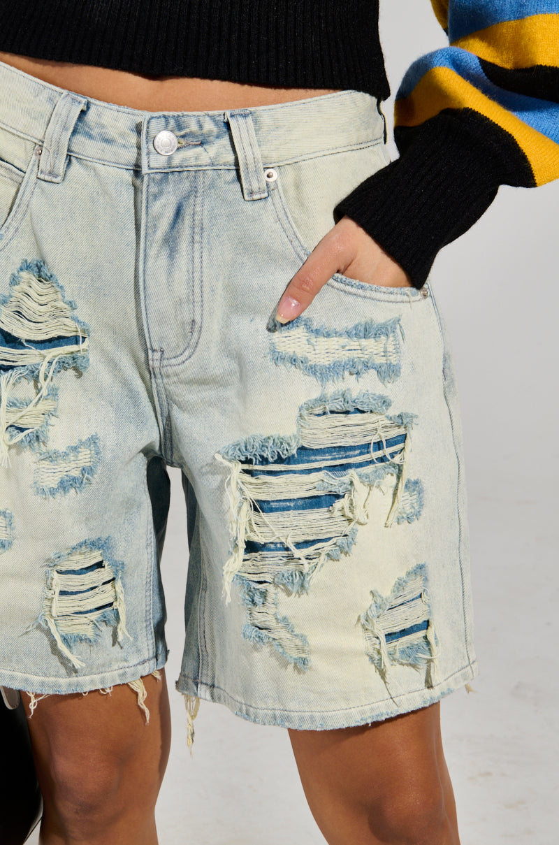 A person wearing the LITE UP DENIM SHORT, featuring a light wash and high-rise fit with distressed detailing, stands with a hand in the pocket. The black top is accented by yellow and blue stripes on the sleeve against a plain, light-colored background.