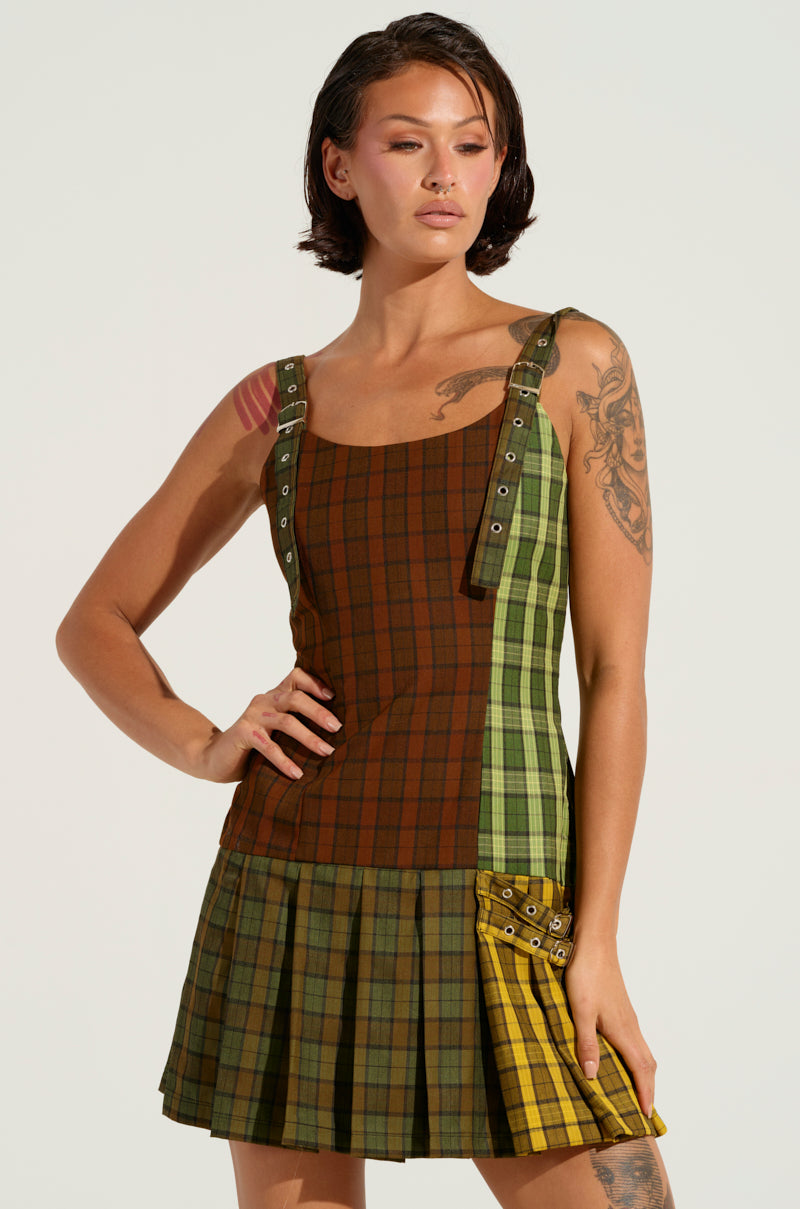 A woman wearing the PLAYTIME IS OVER PLAID MINI DRESS, which showcases an array of green and brown patterns. The dress features adjustable belt straps, a pleated flared skirt, and a yellow plaid accent pocket. She is styled with short hair and has visible tattoos on her arm while posing confidently with her hand on her hip against a plain background.