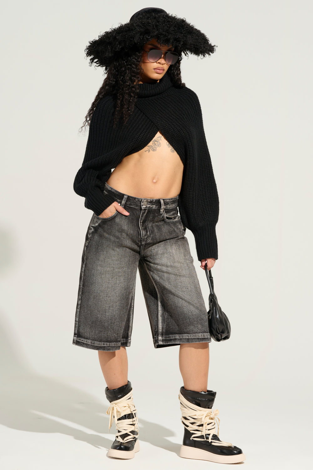 A person with long curly hair is adorned in a large black hat, sunglasses, a black cropped sweater, and high-top sneakers featuring crisscross laces. For added style, they choose the LITERAL PERFECTION BLACK DENIM BERMUDA SHORTS in a baggy fit that exudes confidence while holding a small black purse against a light background. 3
