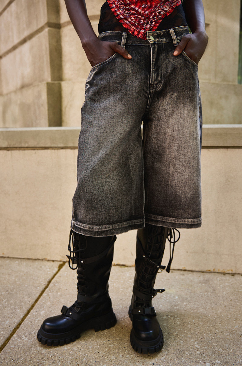 Someone stands with hands in pockets on a concrete sidewalk in front of a stone building, wearing LITERAL PERFECTION BLACK DENIM BERMUDA SHORTS with a baggy fit, paired with a red patterned top and black lace-up combat boots.