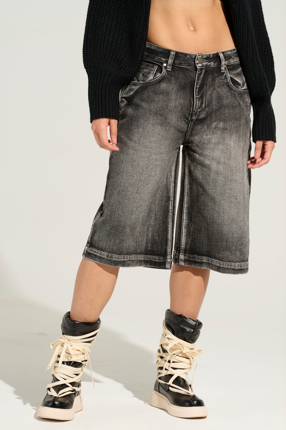 A person wearing a black knit sweater and LITERAL PERFECTION BLACK DENIM BERMUDA SHORTS stands against a light gray background. Chunky black and white high-top sneakers with exaggerated cream laces complete the look. 2