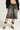 A person wearing a black knit sweater and LITERAL PERFECTION BLACK DENIM BERMUDA SHORTS stands against a light gray background. Chunky black and white high-top sneakers with exaggerated cream laces complete the look.