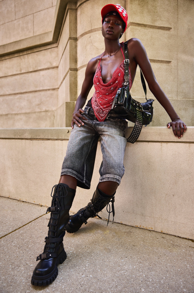 A person in a red beaded top, LITERAL PERFECTION BLACK DENIM BERMUDA SHORTS, black lace-up boots, a red cap, and a black crossbody bag sits confidently on a stone ledge against a beige brick wall. 1