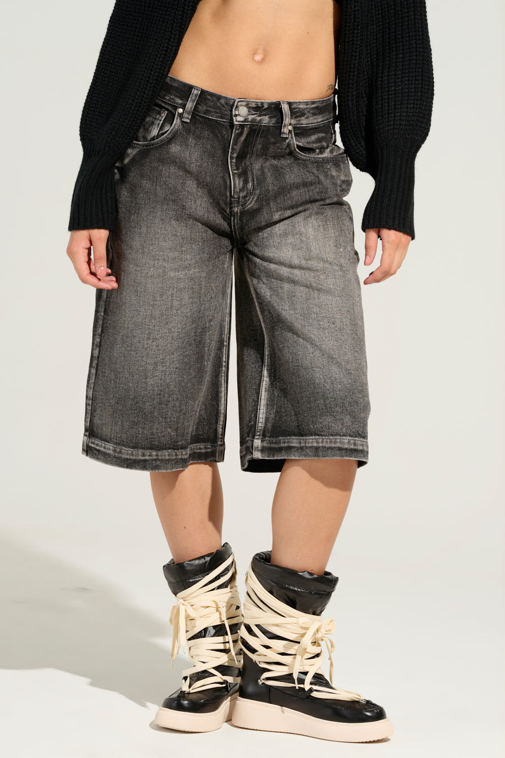 A person stands against a plain background wearing LITERAL PERFECTION BLACK DENIM BERMUDA SHORTS, characterized by their baggy fit, paired with a black sweater and black boots featuring thick beige laces. The lower half of their outfit draws the viewer's focus. 4