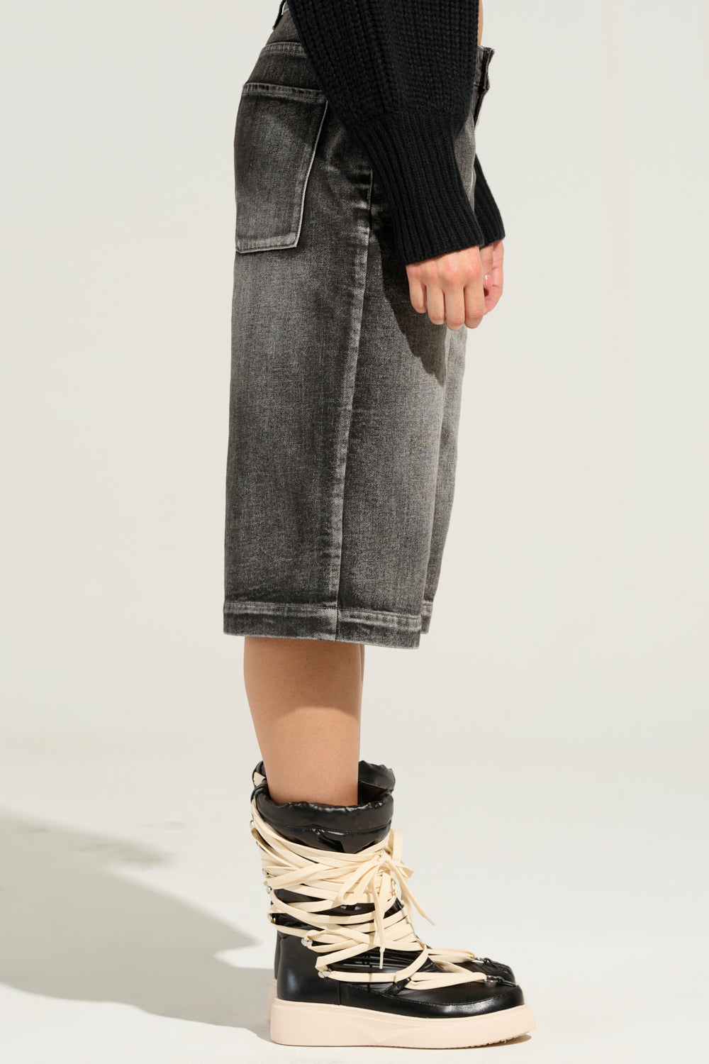 A person is dressed in a black sweater and high-top sneakers with white straps. The side view reveals an outfit featuring the LITERAL PERFECTION BLACK DENIM BERMUDA SHORTS, knee-length with a baggy fit and elements of a high-rise design, all set against a plain background. 5