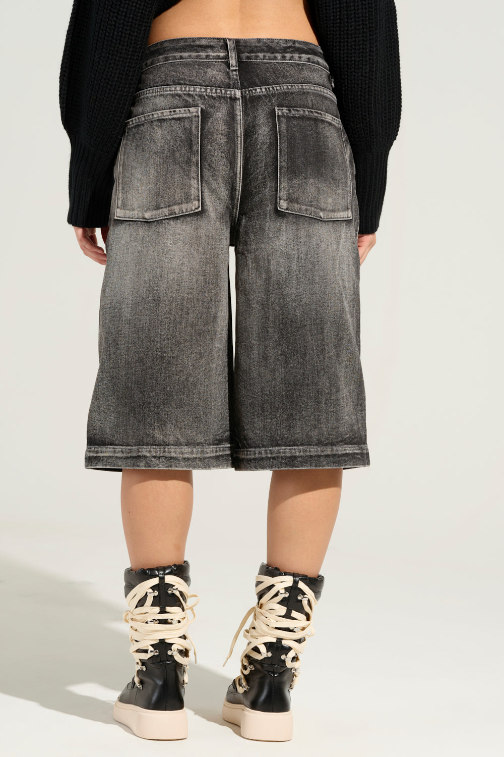 A person sporting the LITERAL PERFECTION black denim Bermuda shorts stands with their back to the camera. Complementing their look, they wear a black sweater and high-top sneakers with thick white laces, effortlessly blending comfort and style. 6