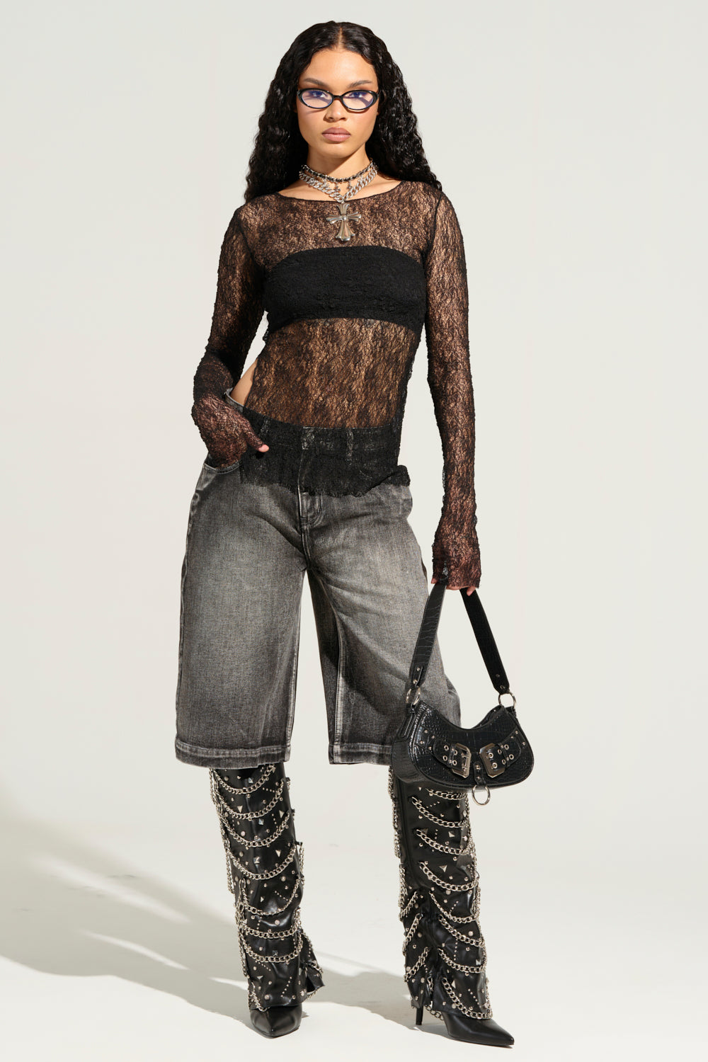 A person is wearing a sheer black lace top paired with LITERAL PERFECTION BLACK DENIM BERMUDA SHORTS. They complement the look with tall black boots adorned with chains, holding a small black handbag while sporting glasses. The scene is set against a plain, light-colored background. 7