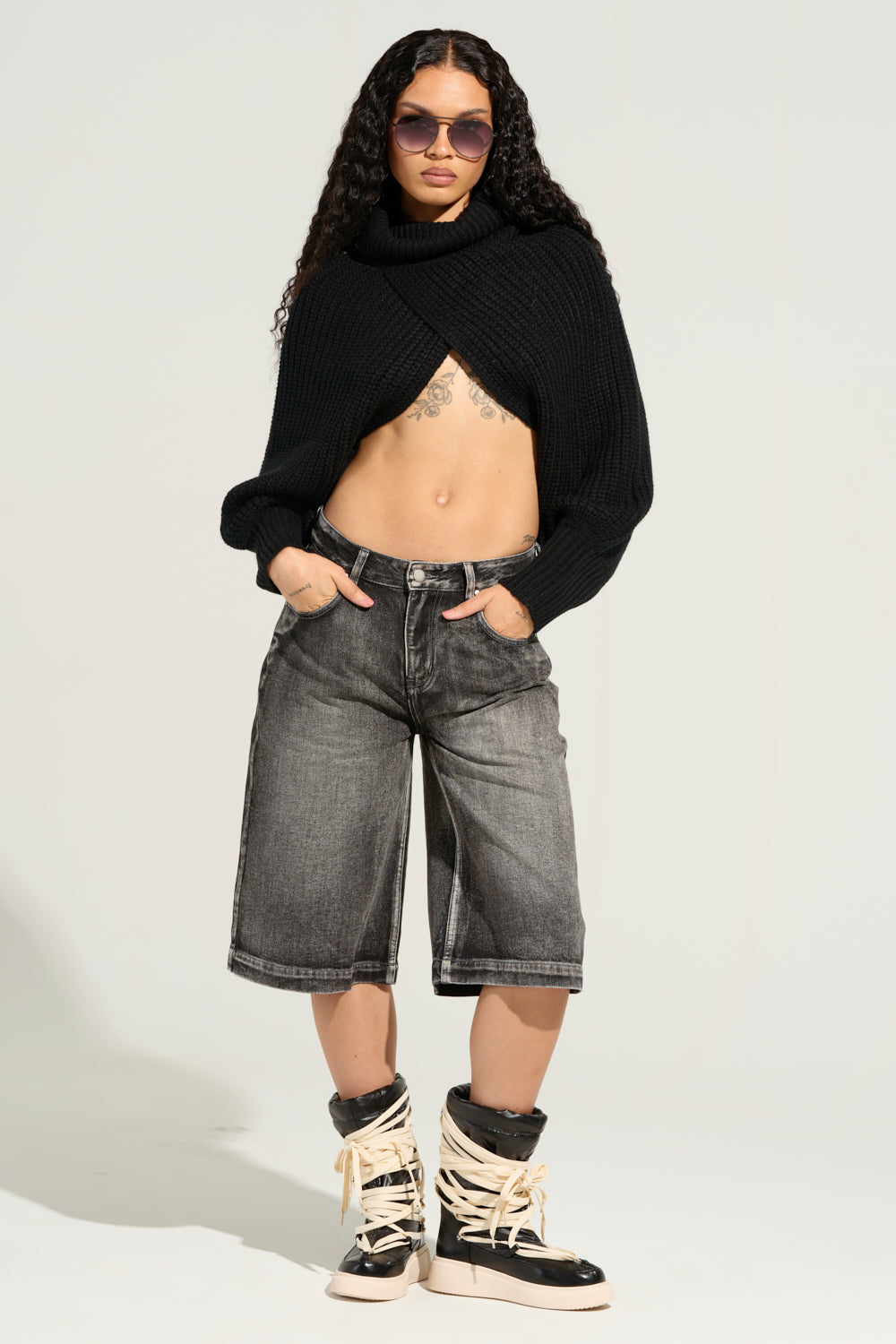 A person with long curly hair stands confidently, wearing large sunglasses, a black cropped sweater, and the LITERAL PERFECTION BLACK DENIM BERMUDA SHORTS. Their high-top boots with lace straps add flair to the baggy fit ensemble. With hands in pockets against a neutral background, they embody a cool, high-rise design aesthetic. 8