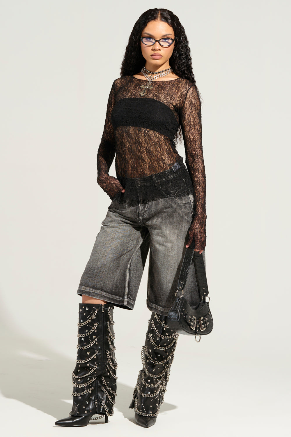 A person in a black lace top and LITERAL PERFECTION BLACK DENIM BERMUDA SHORTS stands confidently, adorned with long, curly hair, glasses, and accessories including necklaces and chain-detailed boots. The ensemble is completed by a baggy fit black handbag. 9