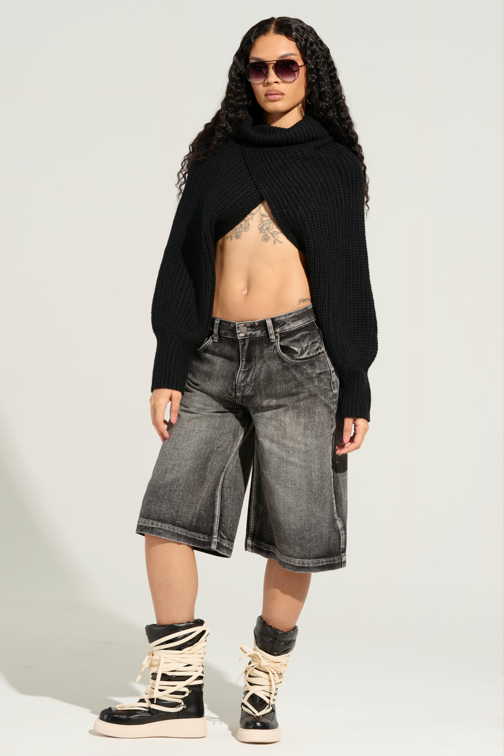 A person with long curly hair and sunglasses confidently stands against a light background, wearing a black cropped sweater and the LITERAL PERFECTION BLACK DENIM BERMUDA SHORTS featuring a high-rise design and baggy fit. Their look is completed with black high-top boots adorned with beige laces, revealing tattoos on their torso. 10