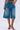 A person in LITERAL PERFECTION BLUE DENIM BERMUDA SHORTS—baggy fit, knee-length, frayed hem, high rise—paired with heeled sandals and one hand in a pocket, stands against a plain light gray background.