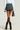 A person with dark skin is wearing a white long-sleeved shirt, a green and blue BON VOYAGE STRIPED DENIM PLEATED MINI SKIRT, sheer black thigh-high stockings, and black heeled boots. They are holding a small black handbag and standing against a light-colored background.