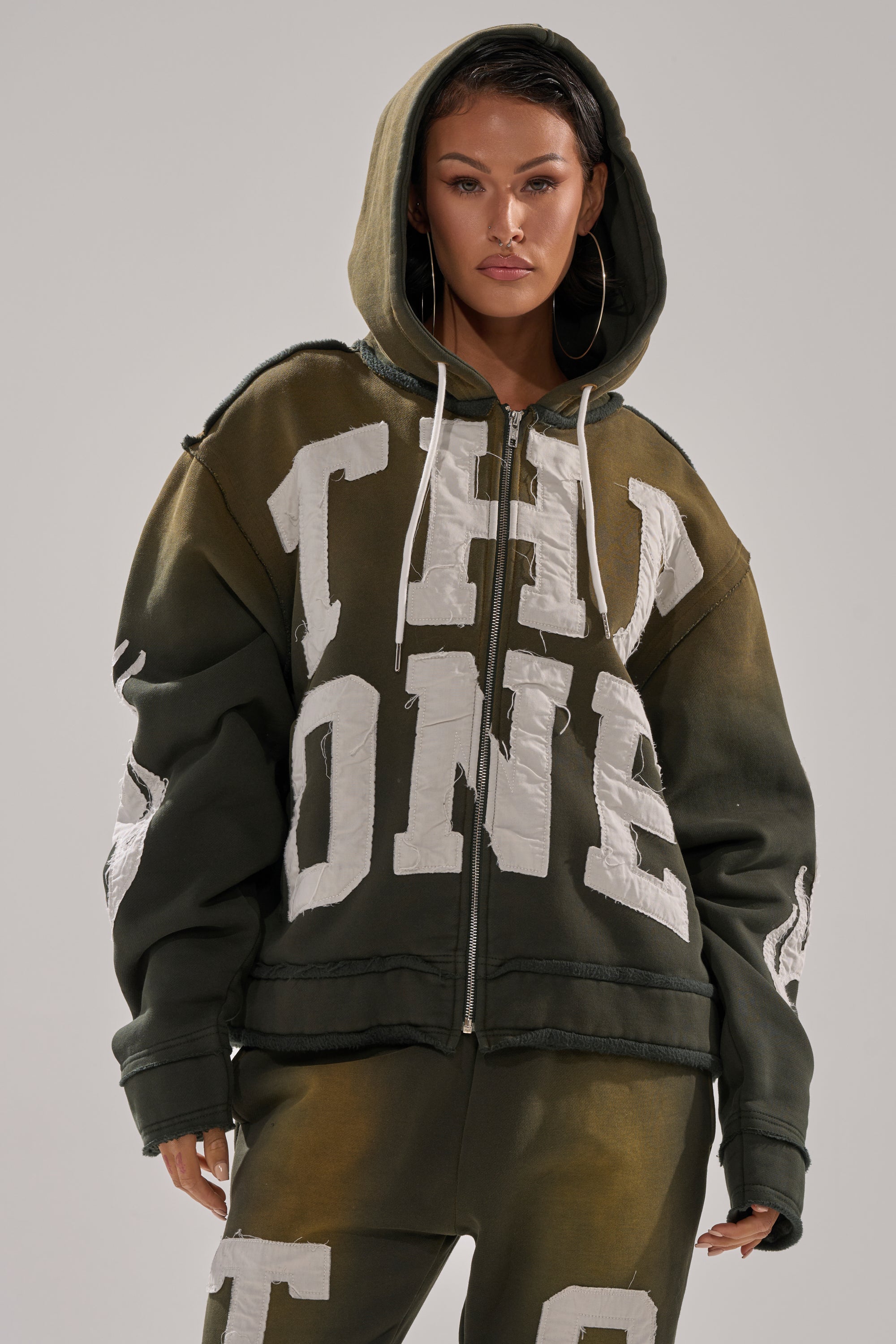 A woman faces forward in THE ONLY ONE ZIP SWEATSHIRT, an oversized green zip hoodie with bold white block letters. She wears the hood up, large hoop earrings, and has a neutral expression against a plain background. 0