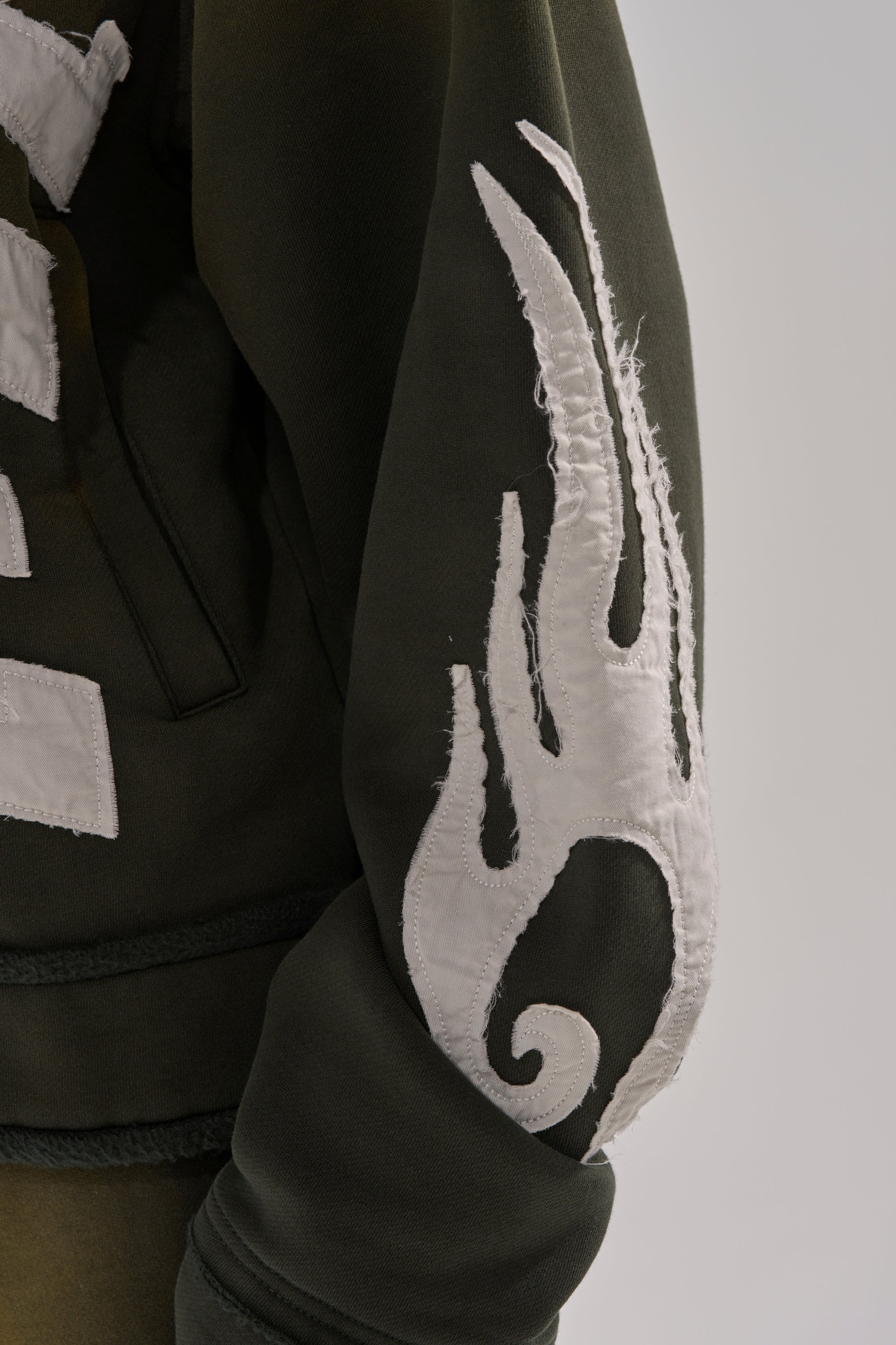 A close-up of THE ONLY ONE ZIP SWEATSHIRT's green oversized sleeve, showcasing a bold white flame-like applique with visible stitching against a neutral background. 9