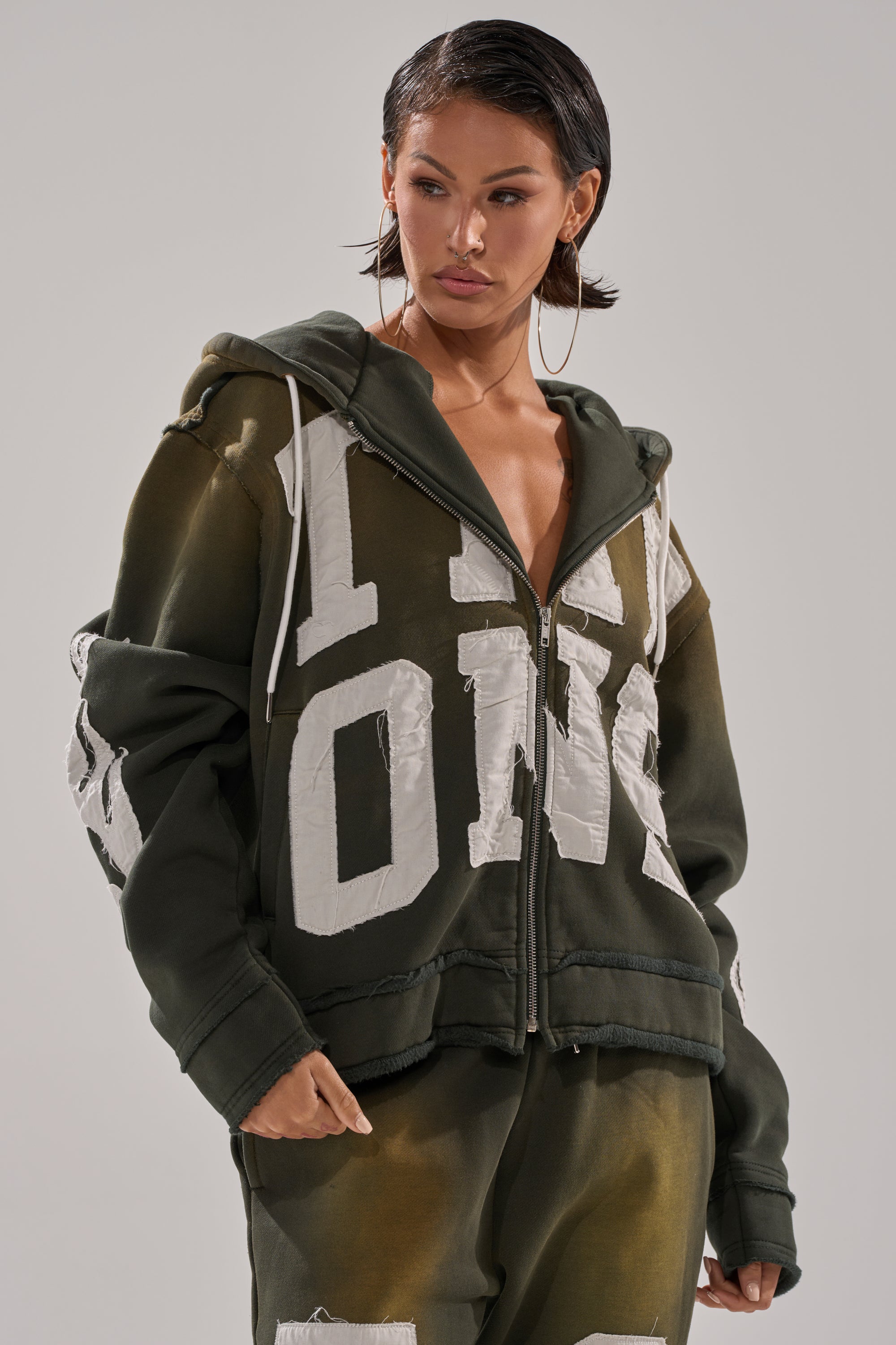 A woman with short, wet-look hair and large hoop earrings wears THE ONLY ONE ZIP SWEATSHIRT in olive green with bold white text and an oversized fit, paired with matching pants, against a plain background. 1