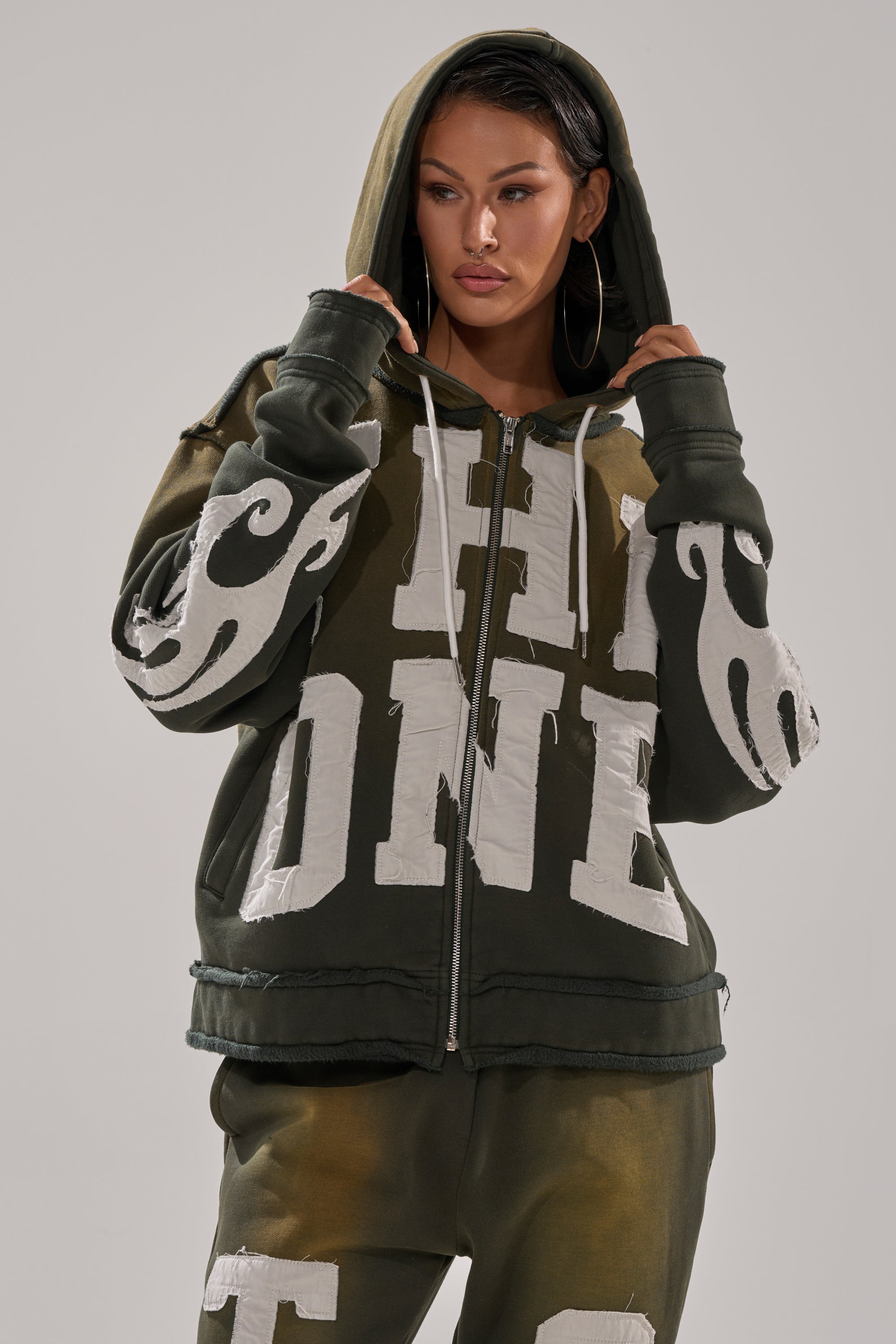 A woman wears THE ONLY ONE ZIP SWEATSHIRT in oversized dark green with bold white lettering and sleeve designs, paired with matching pants. She has the hood up, looks to the side, and holds the sweatshirt near her face. 2