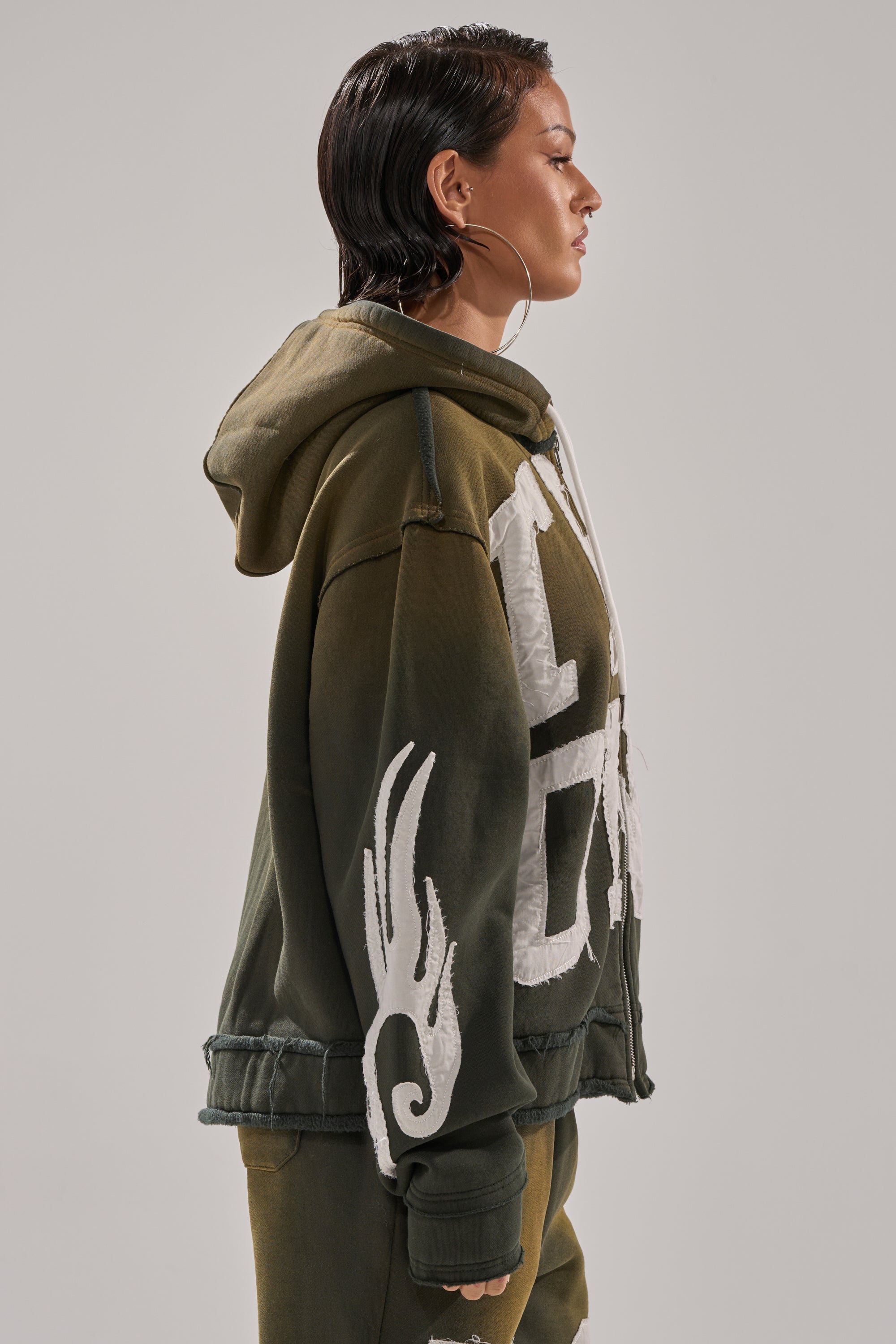 A person with short, wet-look hair and large hoop earrings wears THE ONLY ONE ZIP SWEATSHIRT—an olive-green, oversized zip-up with white graphic designs—in profile against a light gray background. 3