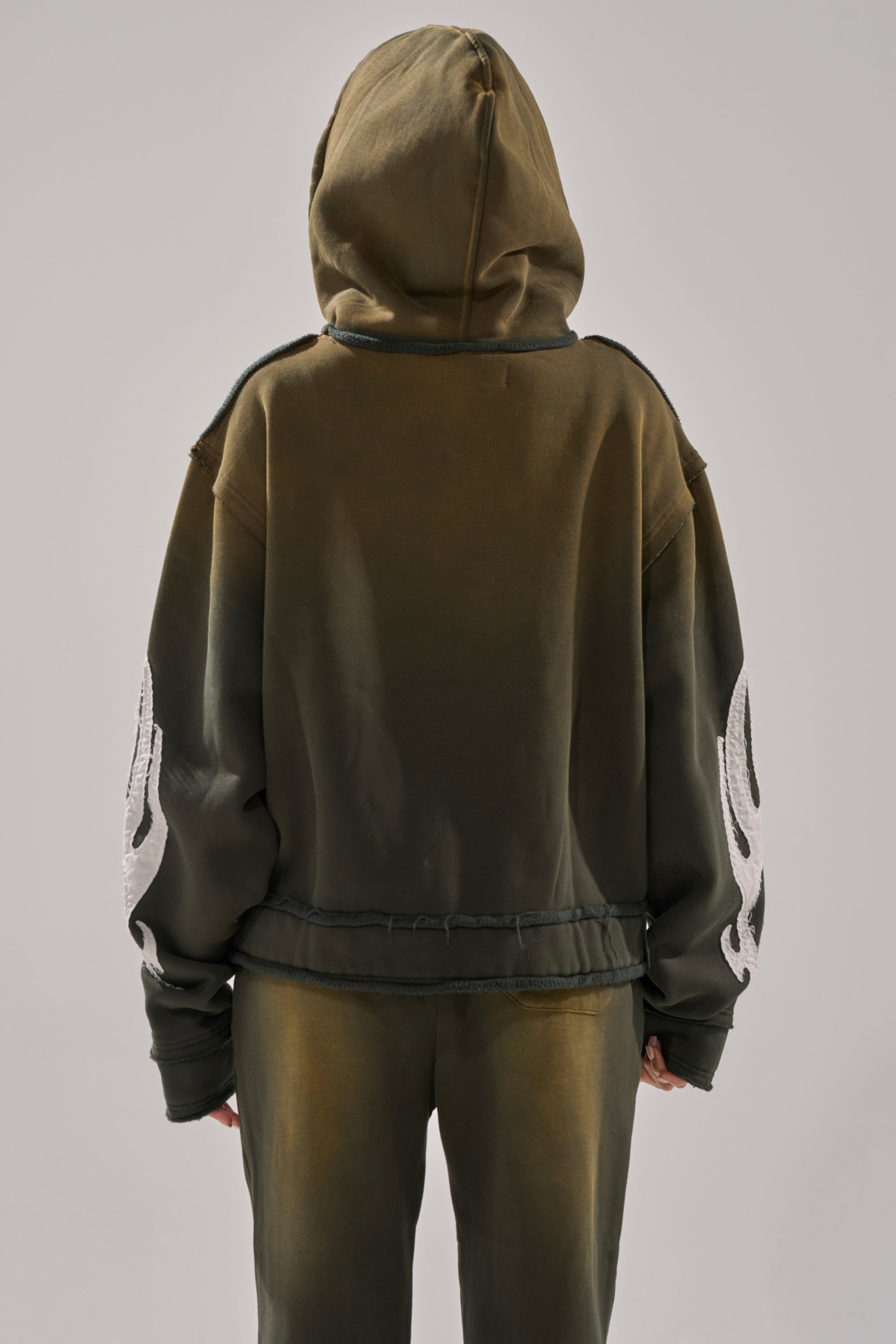 A person faces away from the camera wearing THE ONLY ONE ZIP SWEATSHIRT in olive green, featuring an oversized fit, hood up, and bold white flame designs on the sleeves, paired with matching pants. 4