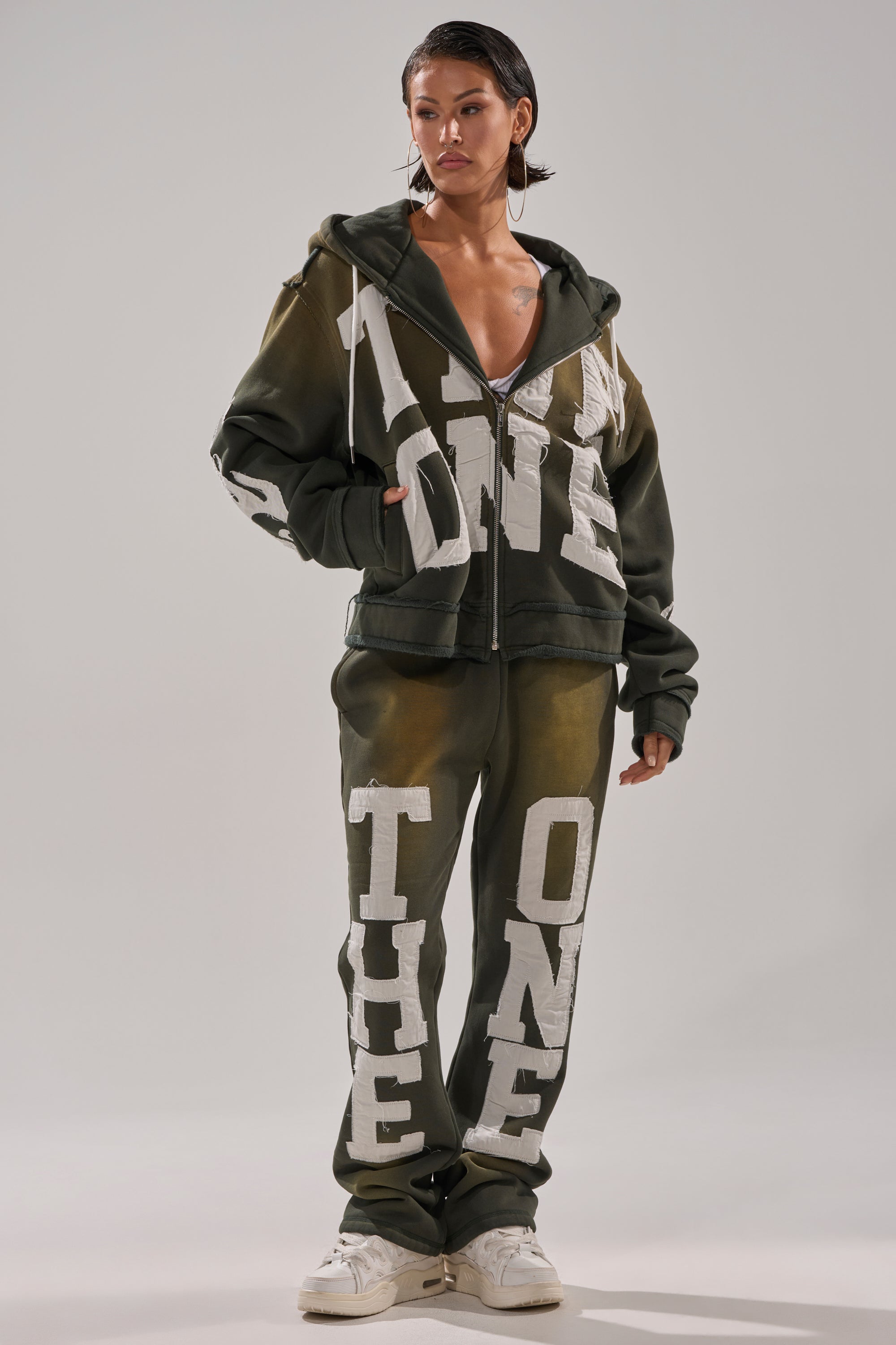 A woman wears an oversized dark green THE ONLY ONE ZIP SWEATSHIRT and matching sweatpants with bold white lettering, styled with white sneakers, wet-look hair, and large hoop earrings. 6