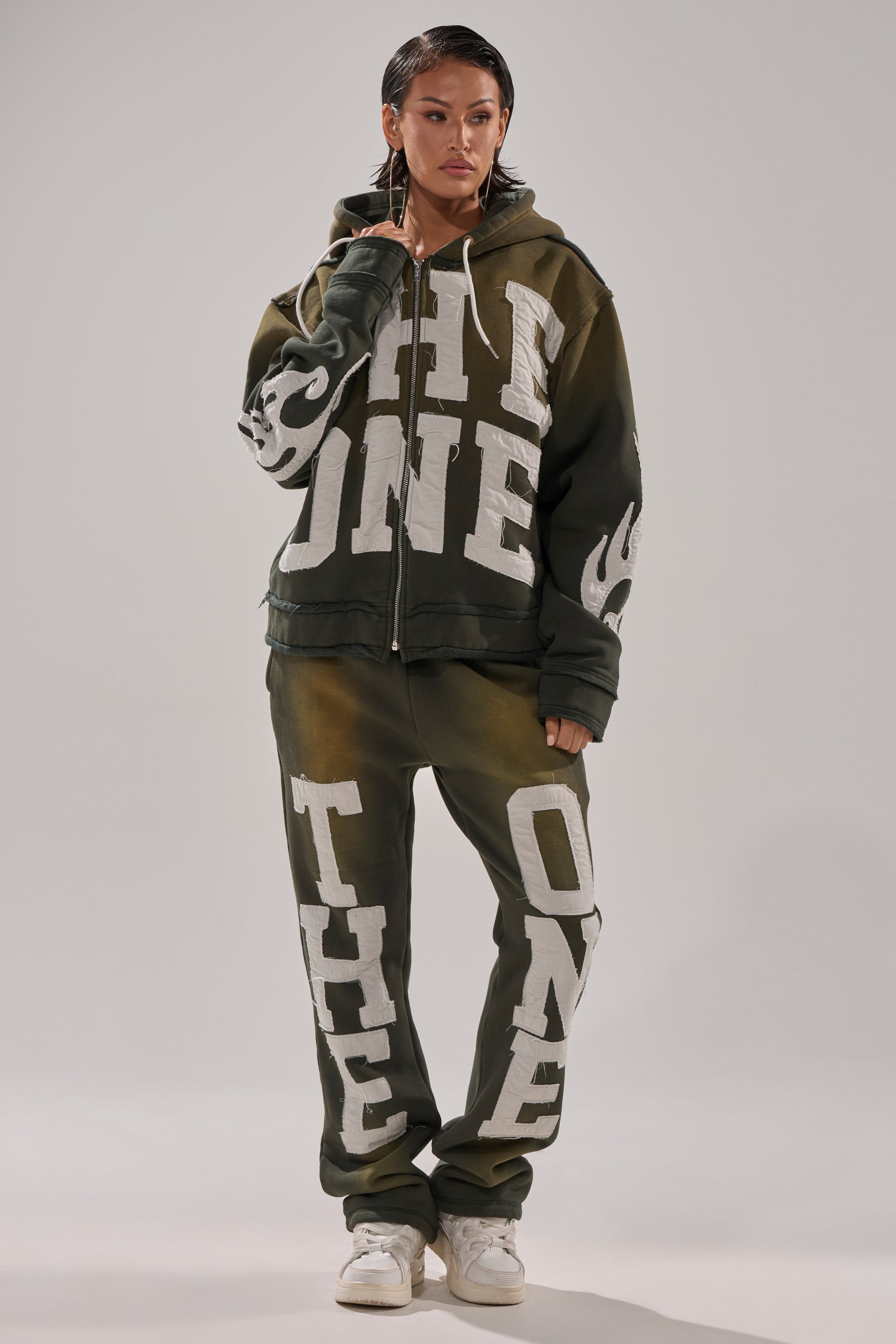 A person wears an oversized olive green THE ONLY ONE ZIP SWEATSHIRT and matching sweatpants with bold white block letters. They style it with wet-look hair, hoop earrings, and white sneakers against a plain light gray background. 7