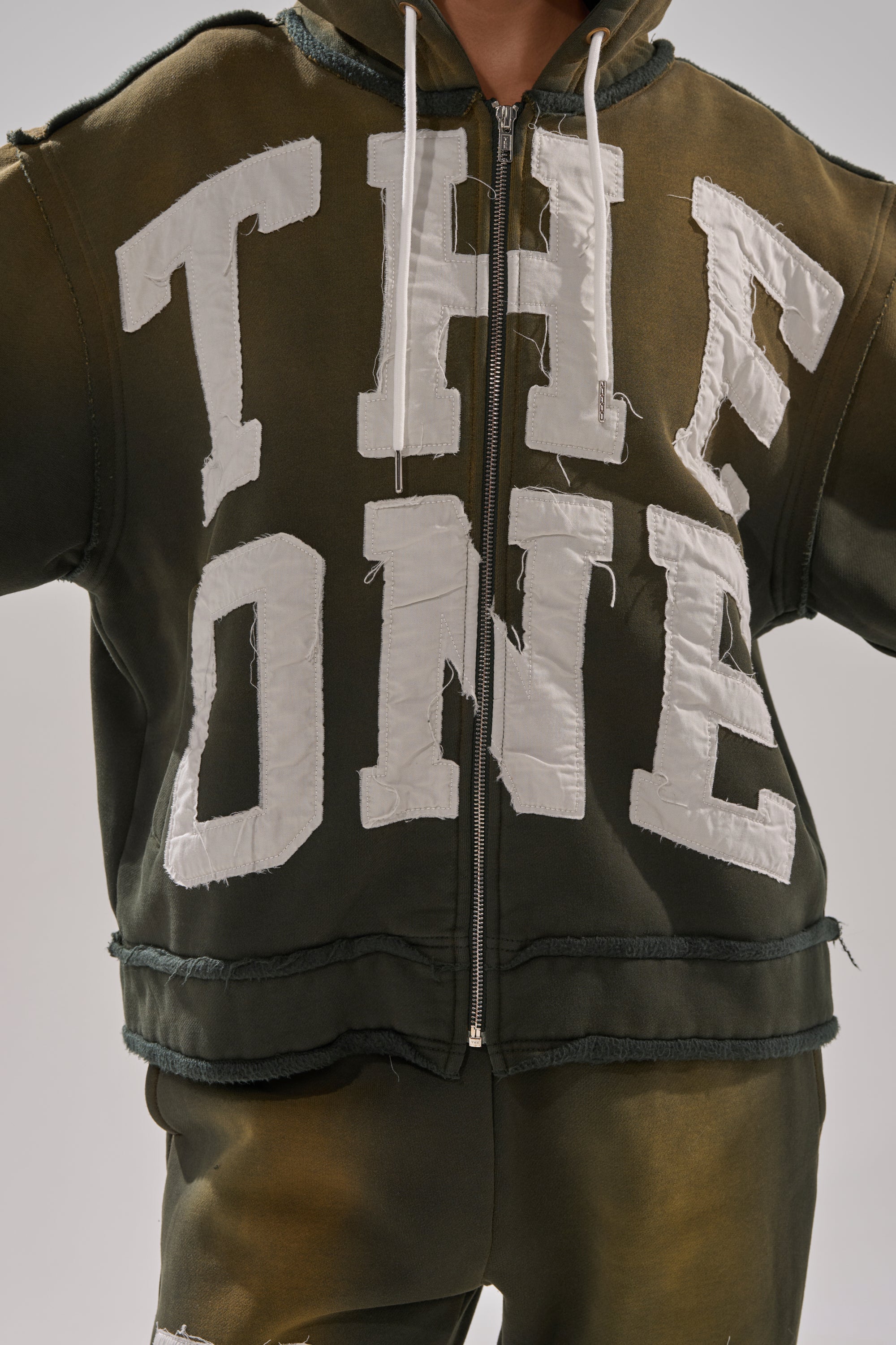A person wears THE ONLY ONE ZIP SWEATSHIRT in dark green with an oversized fit and matching pants. Bold white letters spell "THE ONE" across the front. The image is cropped just below the shoulders and above the knees. 8