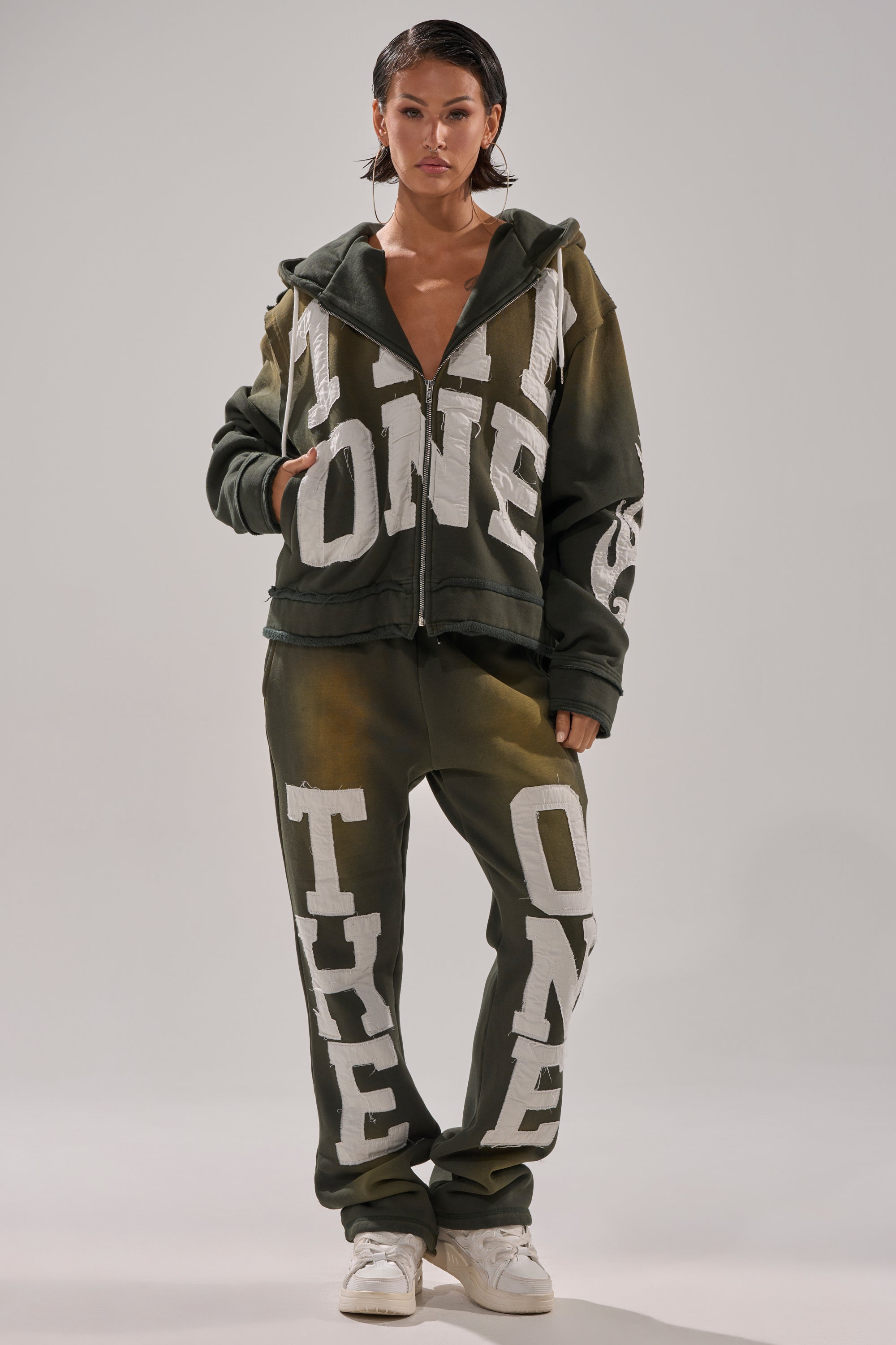 A woman faces forward in an oversized dark green THE ONLY ONE JOGGER set with bold white lettering, wet-look hair, and white sneakers against a plain background. 0