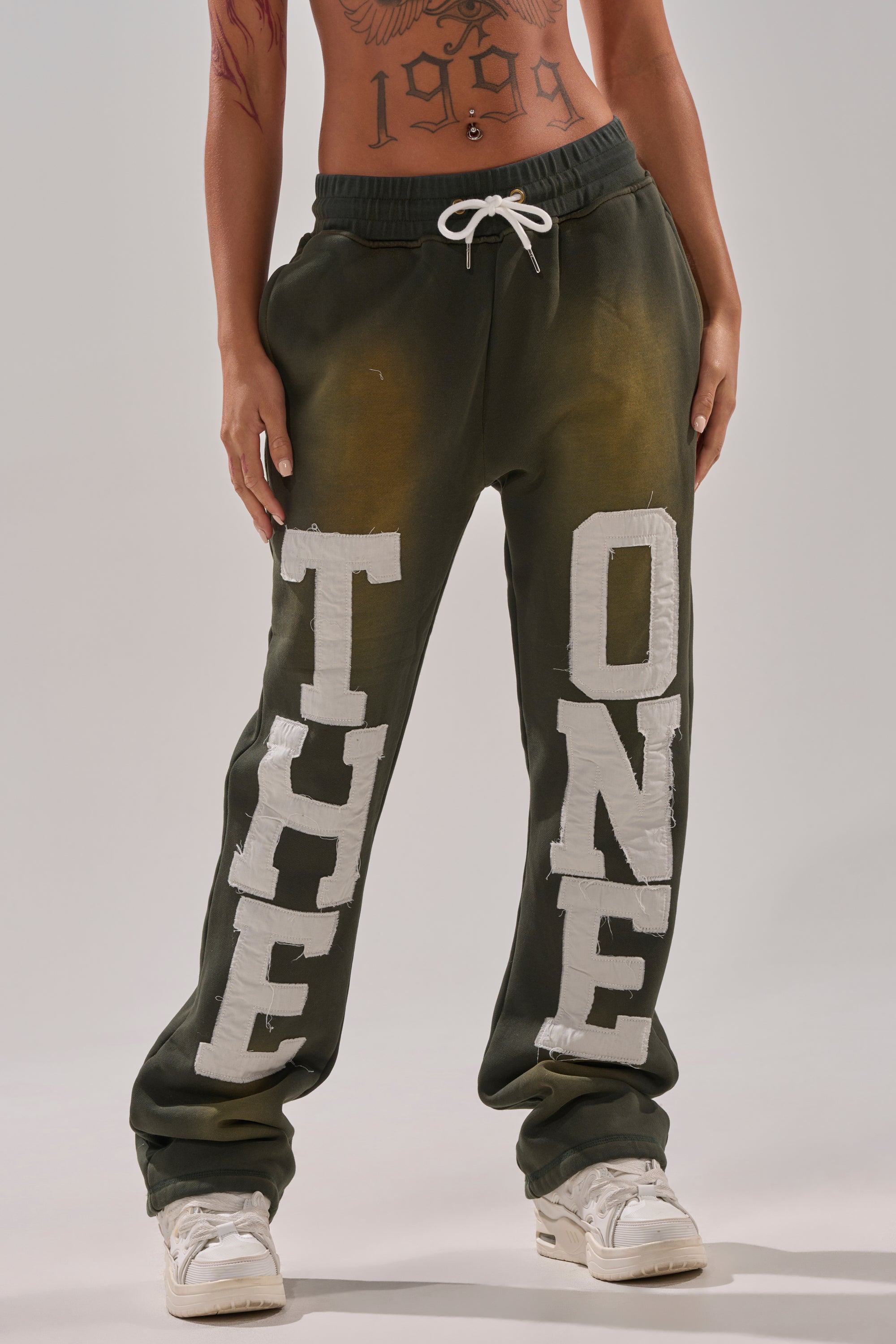A person wears dark green THE ONLY ONE JOGGER sweatpants with large white lettering, paired with white sneakers and displaying a visible torso tattoo; image is cropped from shoulders down. 1