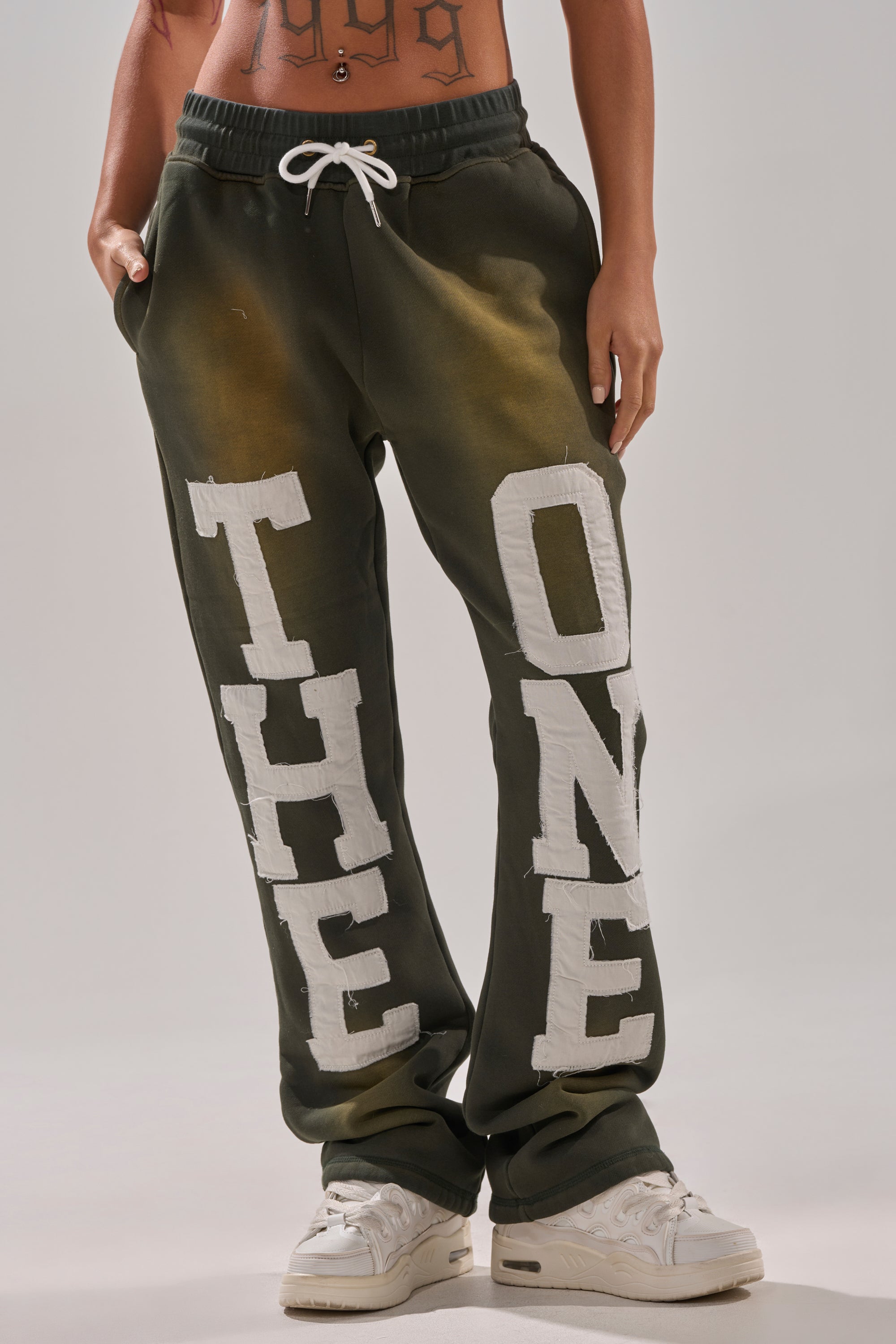 A person wearing dark green THE ONLY ONE JOGGER pants with bold white lettering on the front of each leg, paired with white sneakers. Only the lower torso and legs are visible. 2