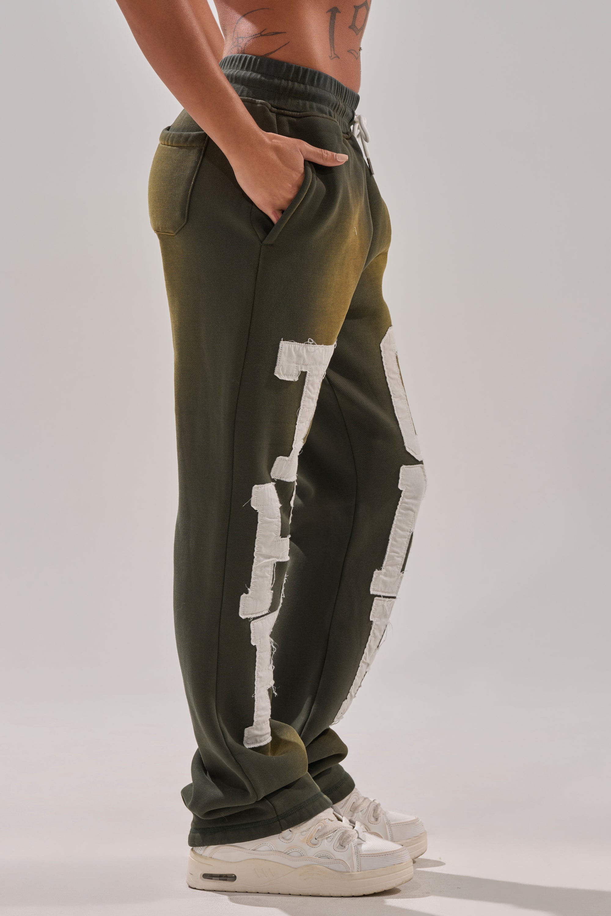 A person stands sideways in THE ONLY ONE JOGGER—olive green cotton-blend sweatpants with large white distressed patches—paired with white sneakers and one hand in their pocket, against a plain light background. 3