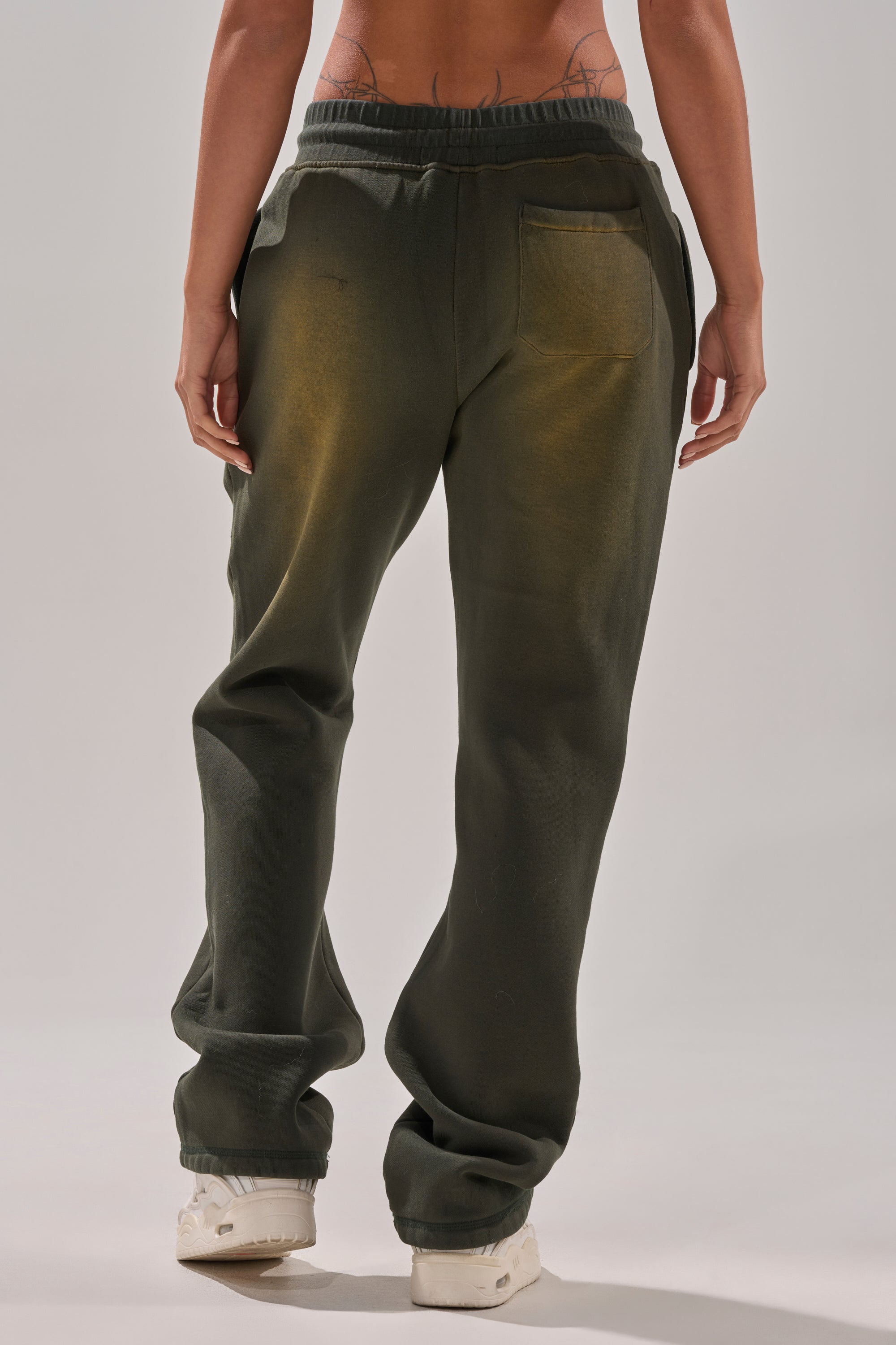 A person faces away, wearing THE ONLY ONE JOGGER in dark green cotton-blend with a back pocket. The loose fit reveals slight yellow stains and partially visible lower back tattoos, paired with white sneakers. 4