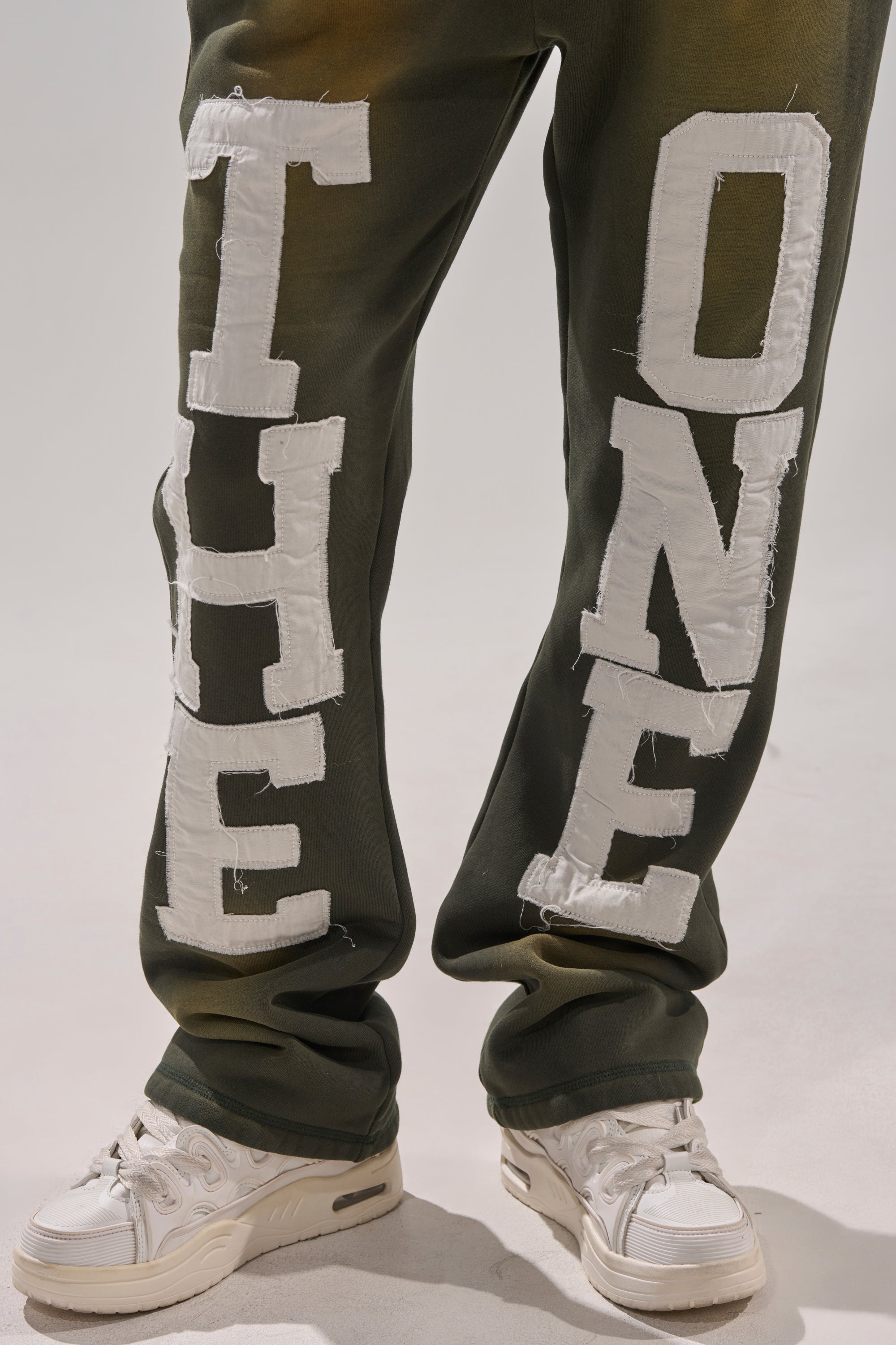 A person wears olive green high-rise THE ONLY ONE JOGGER pants with large white "THE ONLY ONE" lettering down each leg and white sneakers, standing against a plain light background. 7