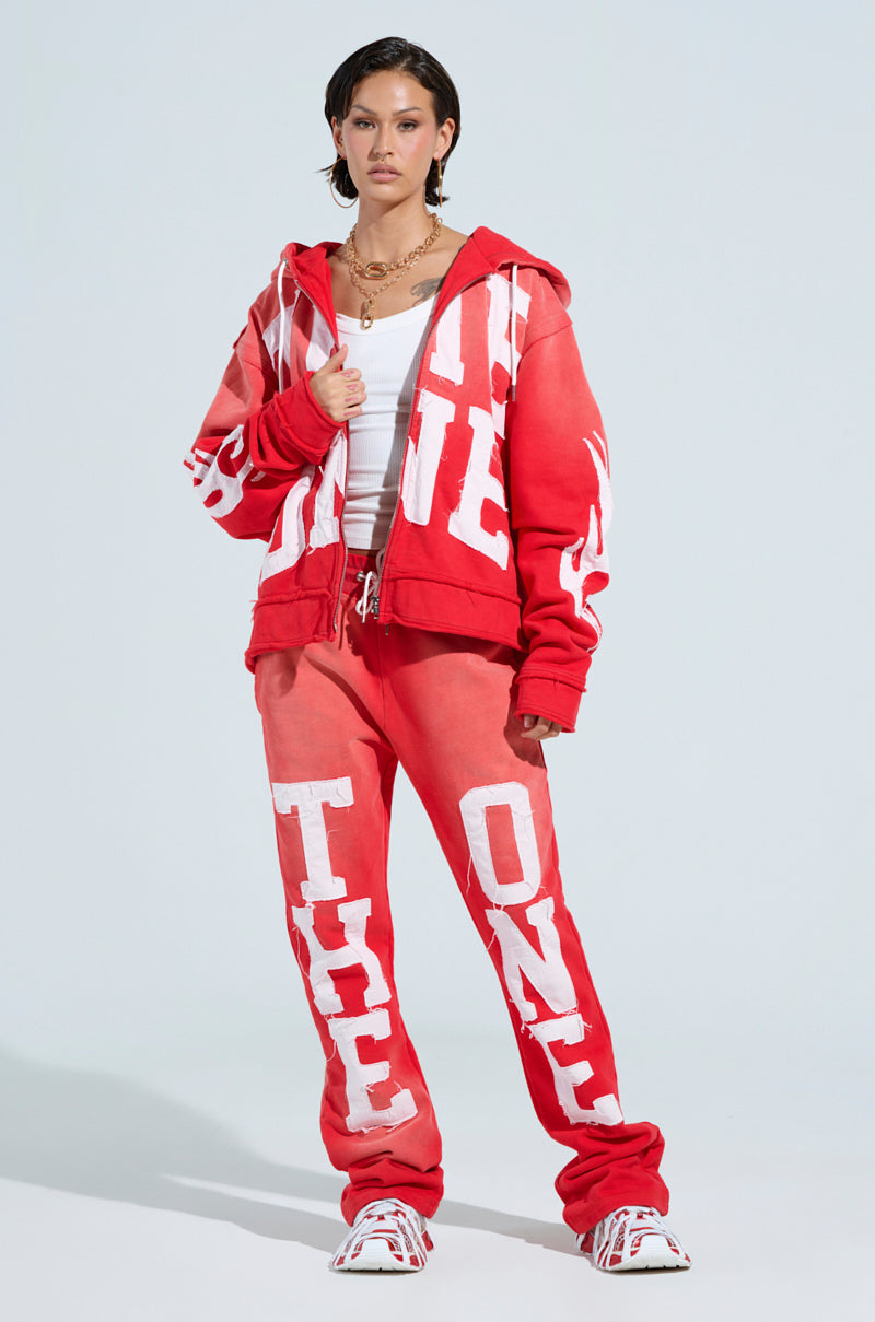A person stands in a red tracksuit with "THE ONE" patches on the ONLY ONE WIDE LEG PANT and jacket, paired with a white tank top. They wear chunky red and white sneakers. The wide-leg pants feature an elastic waistband, all set against a plain light-colored background. 0